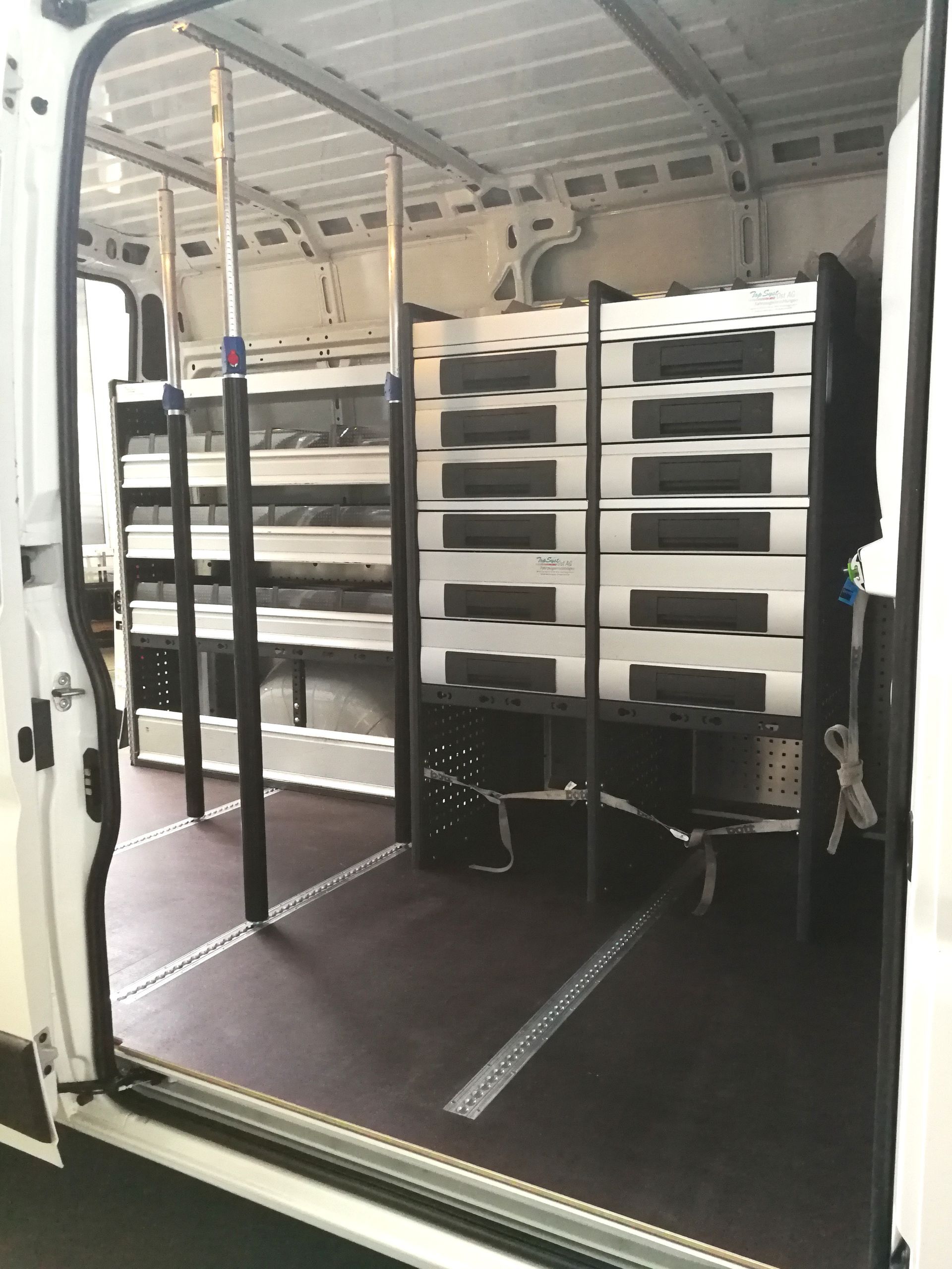 Innenraum eines weißen Lieferwagens mit Regalen, in denen sich graue Kisten befinden, und dunklem Boden.