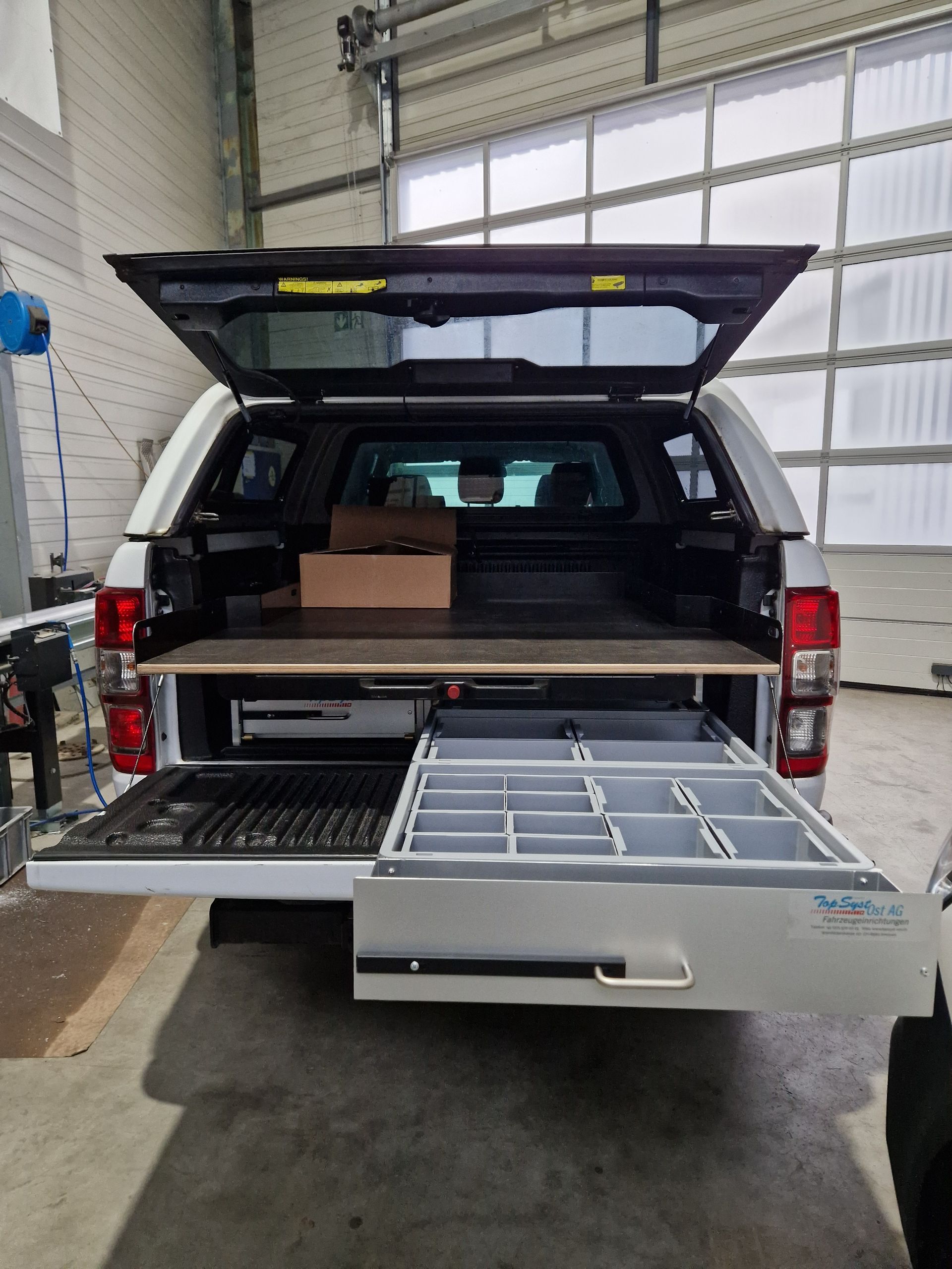 Weiße Ladefläche mit offener Schublade mit Staufächern, schwarze Heckklappe und Dach.