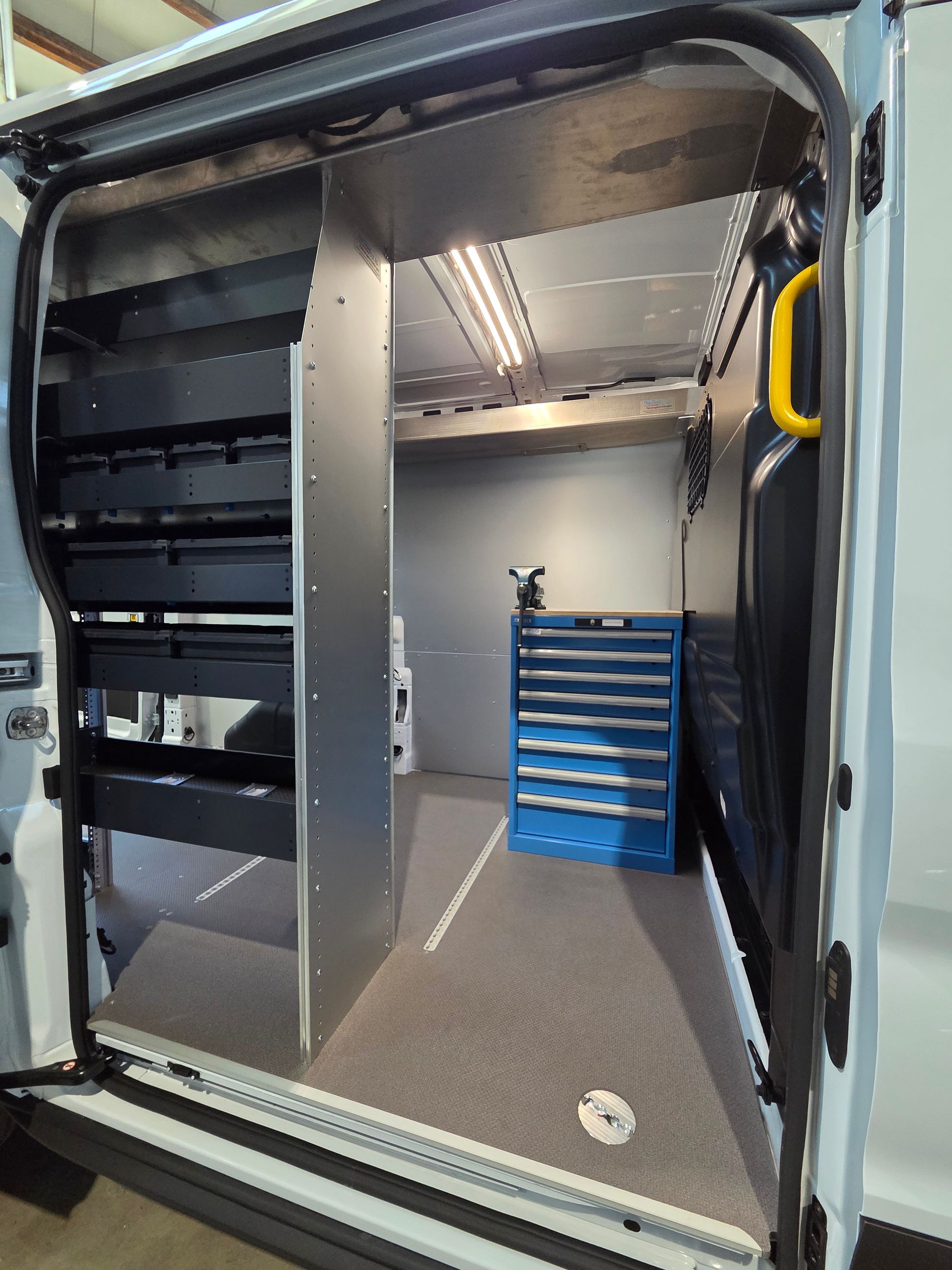 Innenraum eines Arbeitstransporters mit Regalen, einer Werkzeugkiste und einer offenen Tür mit einem gelben Griff.