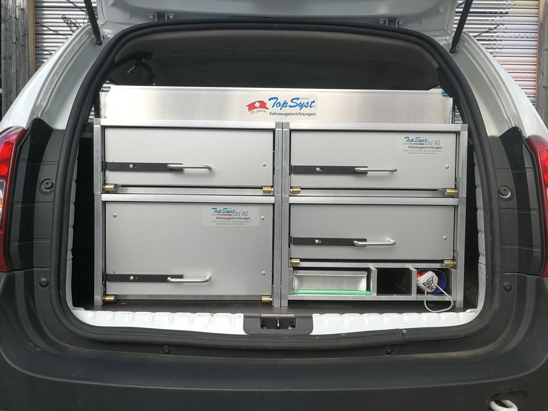Silbernes Aufbewahrungsmöbel im offenen Kofferraum eines weißen Autos mit Schubladen und Fächern.