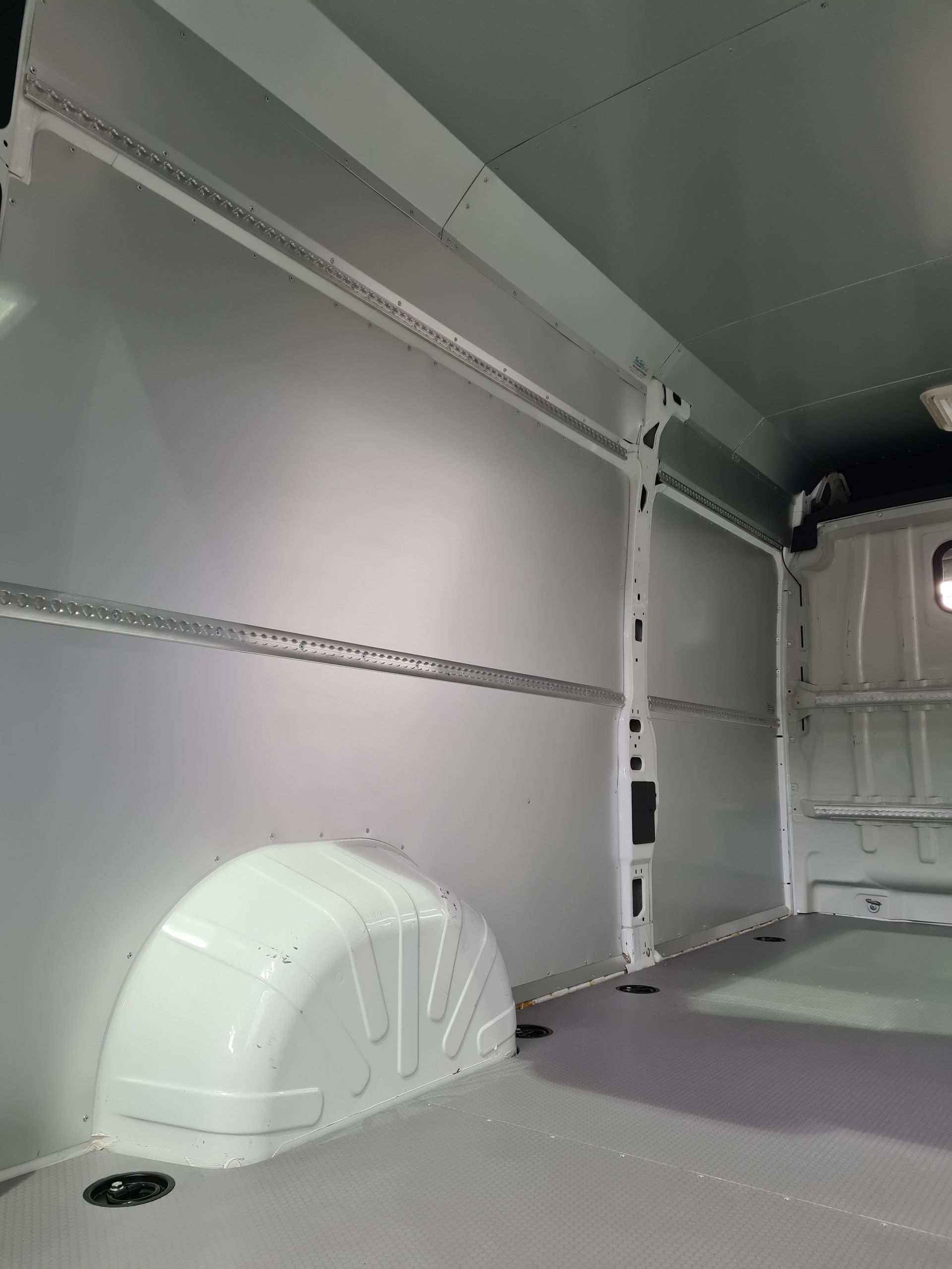 Innenraum eines weißen Lieferwagens mit grauen Wänden und grauem Boden. Metallschienen verlaufen horizontal entlang der Wände.