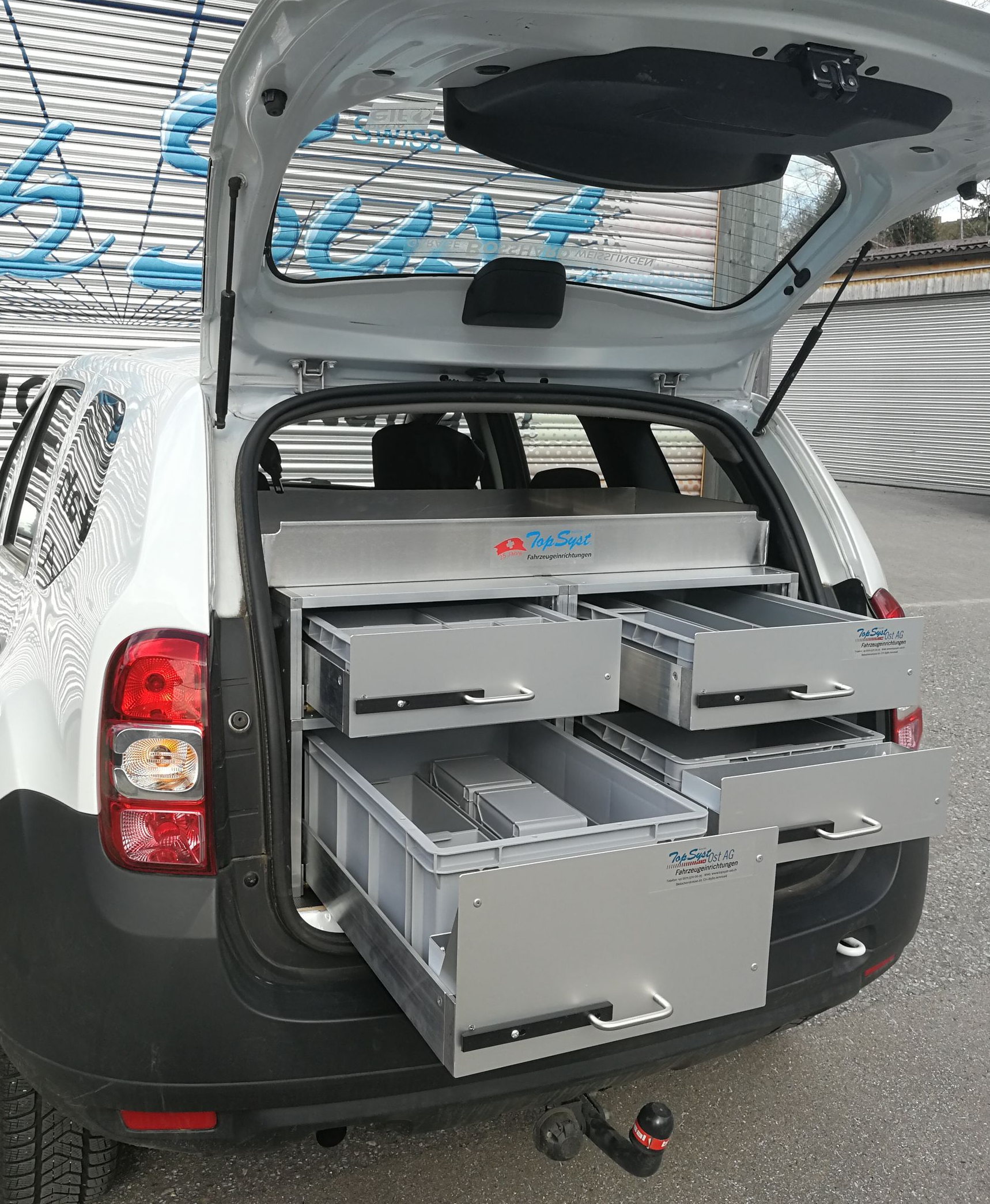 Der Kofferraum eines weißen Autos ist geöffnet und gibt den Blick auf maßgefertigte Schubladen und Ablagefächer aus Metall frei.