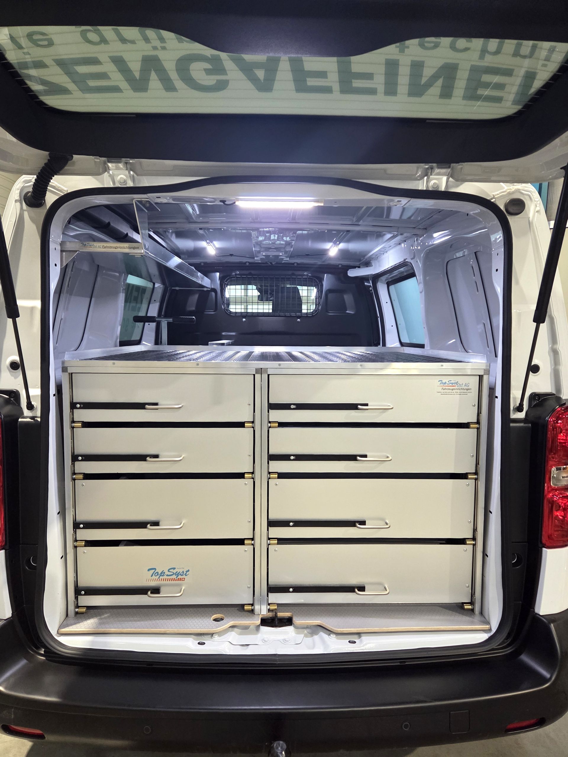Offenes Heck eines weißen Lieferwagens mit Metallschubladen zur Aufbewahrung, beleuchtet durch Deckenleuchten.