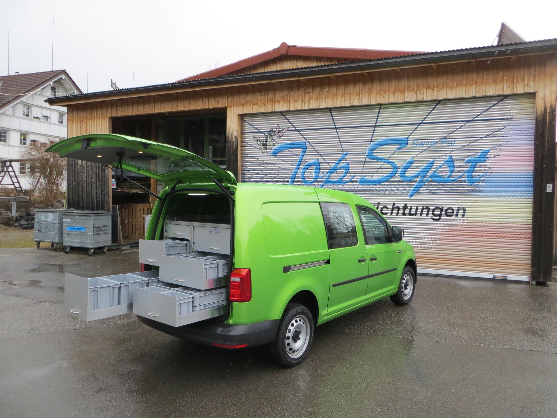 Ein grüner Lieferwagen steht mit geöffnetem Kofferraum vor einer Garage.