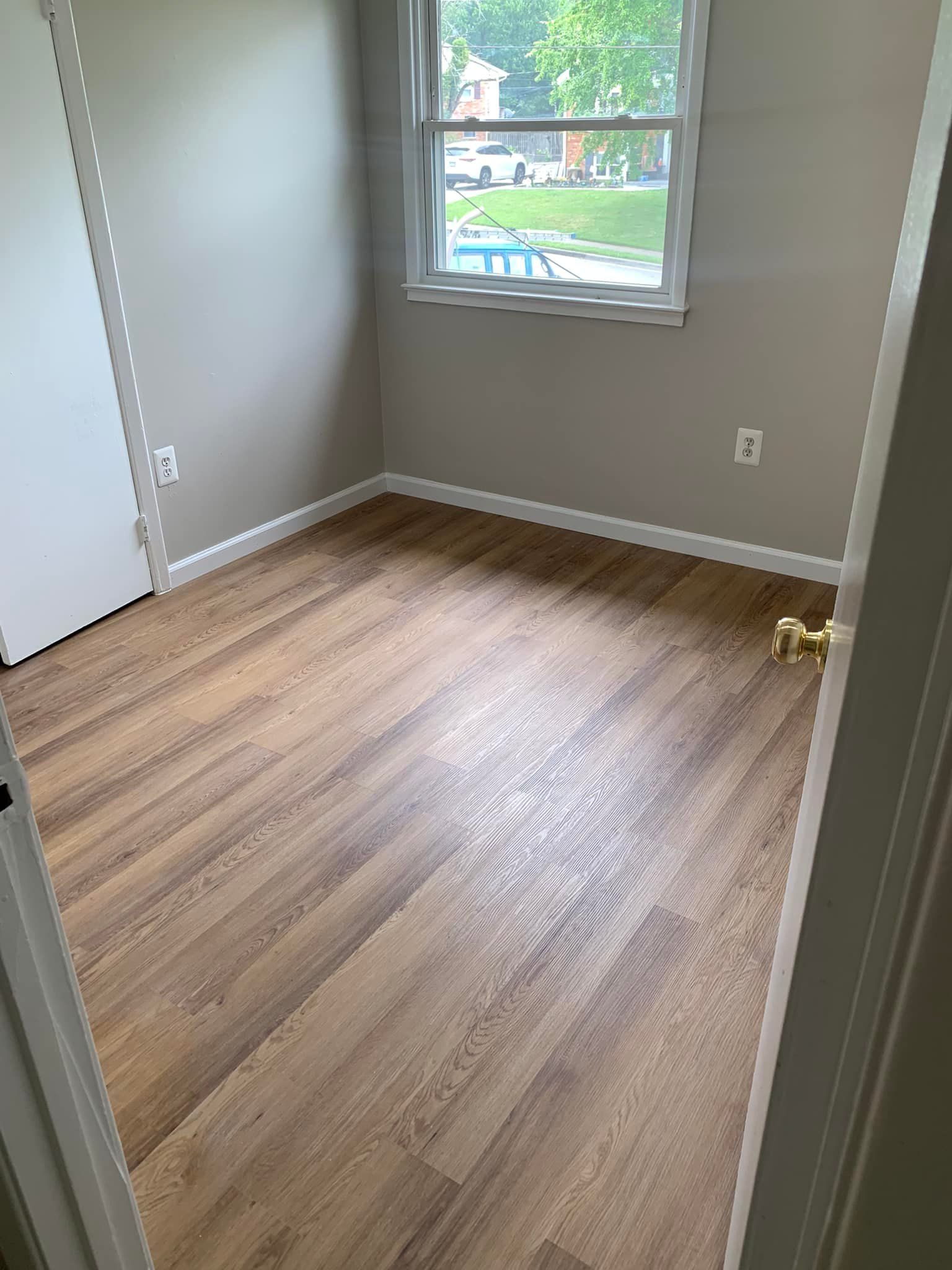 Empty bedroom with wood-look flooring, tan walls, a window, and a white door.
