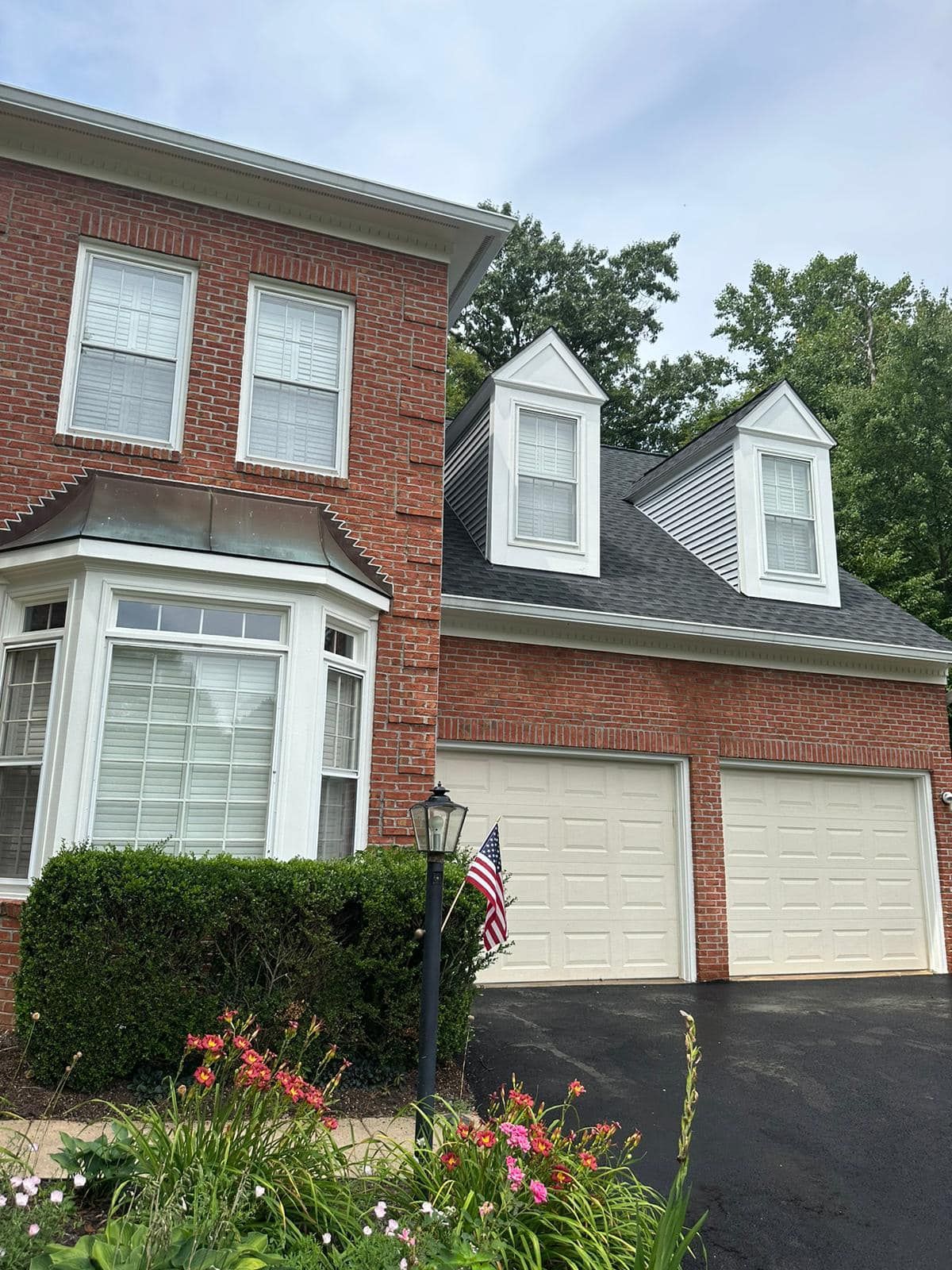 Brick house with a two-car garage, dormers, and a bay window. Landscaping and an American flag are present.