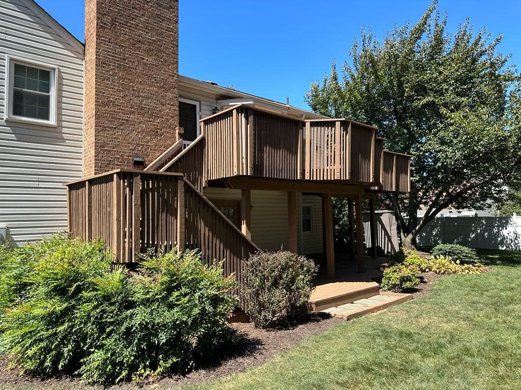 Wooden deck attached to a house with brick chimney, surrounded by green bushes and grass on a sunny day.