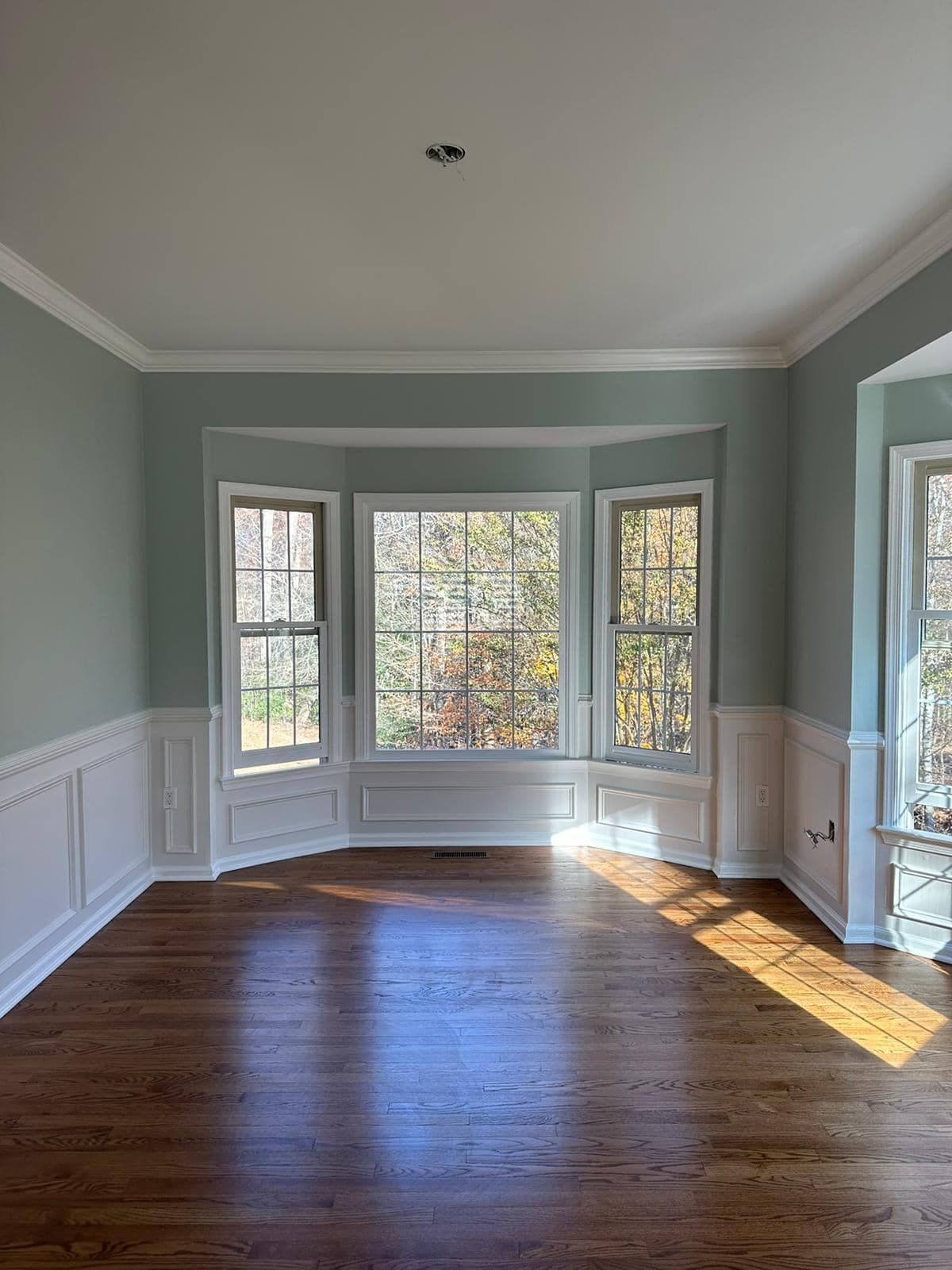 Empty room with hardwood floor, bay window, and teal walls. White trim and wainscoting.