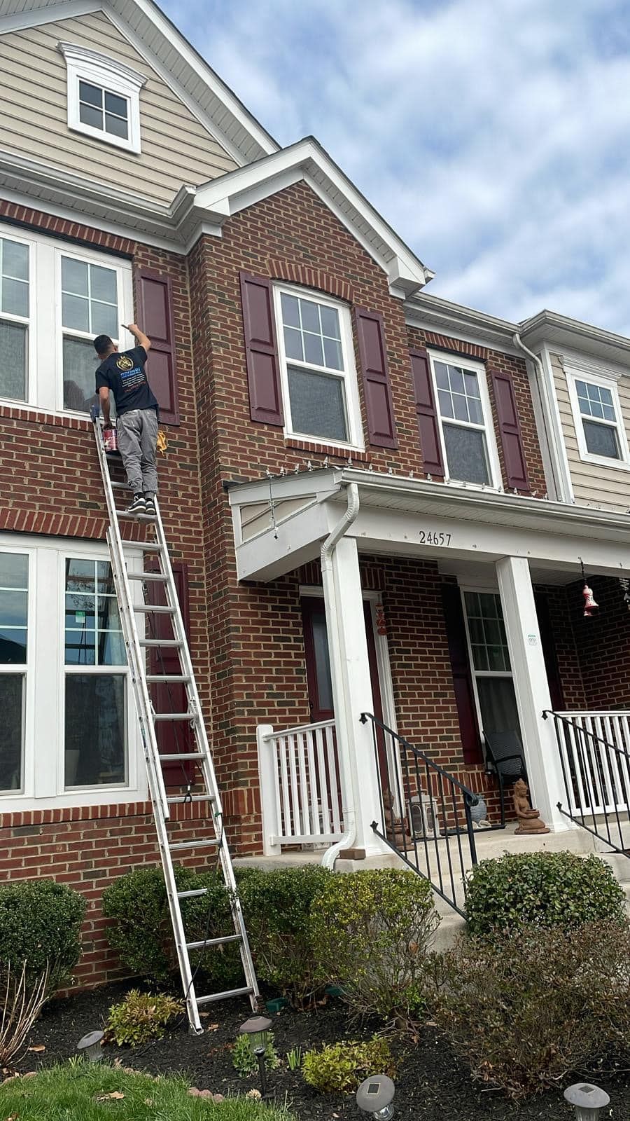 Person on a ladder washing windows of a brick house with maroon shutters. Cloudy sky.
