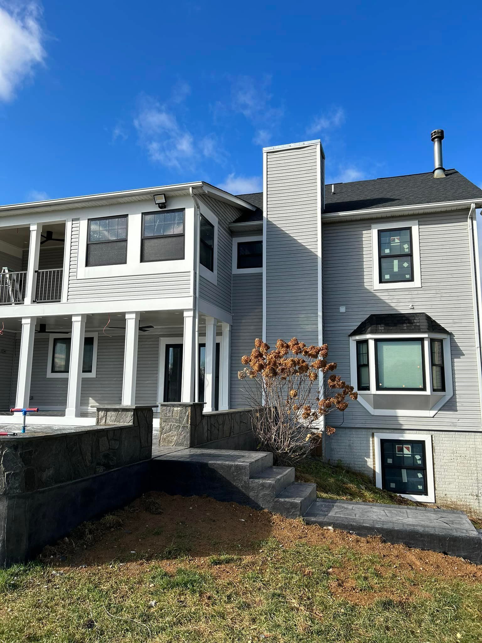 Two-story house with light gray siding, black framed windows, and a brick chimney under a blue sky.