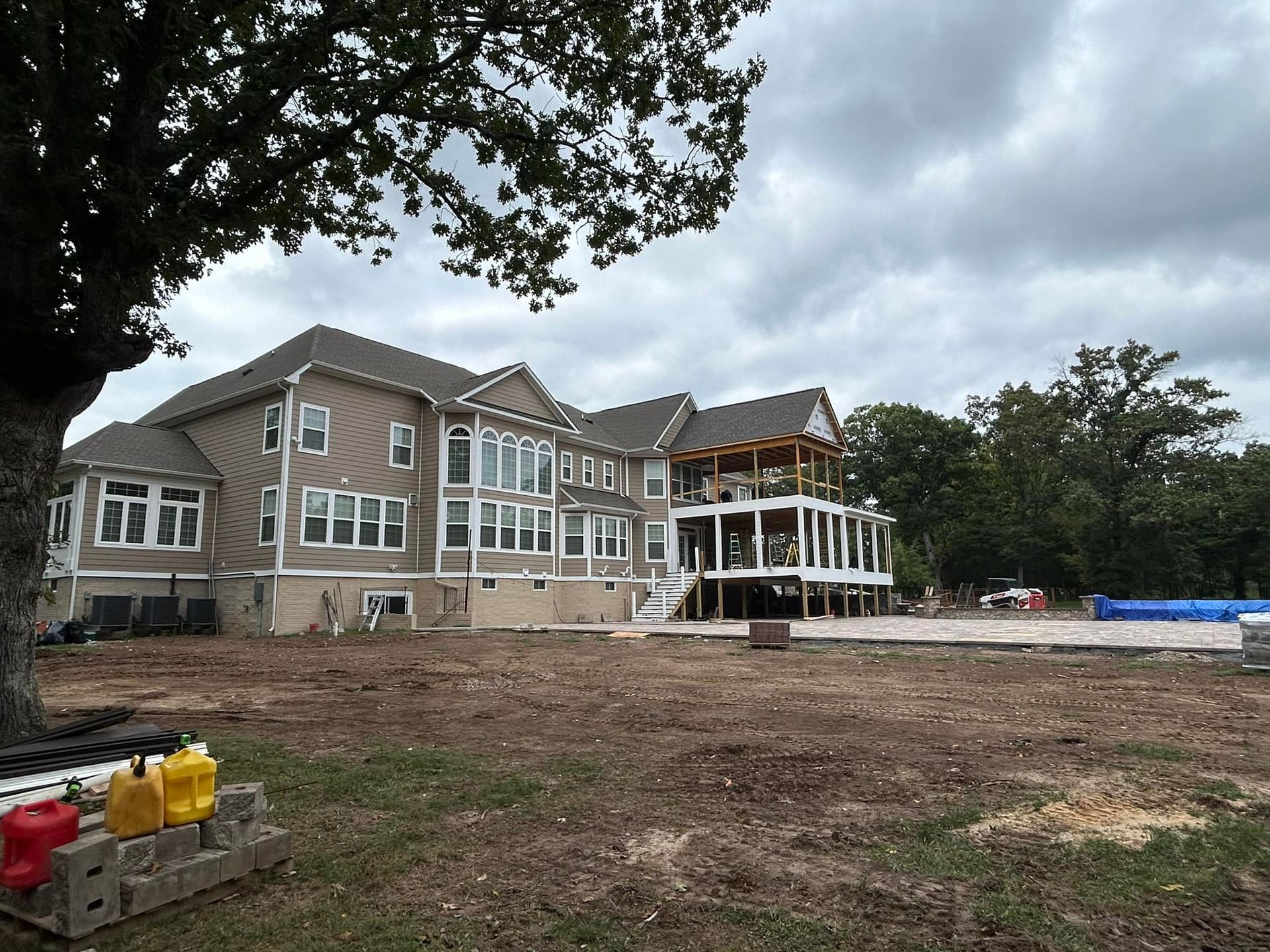 Large house under construction with beige siding, multiple windows, and a covered porch. Cloudy sky and bare yard.