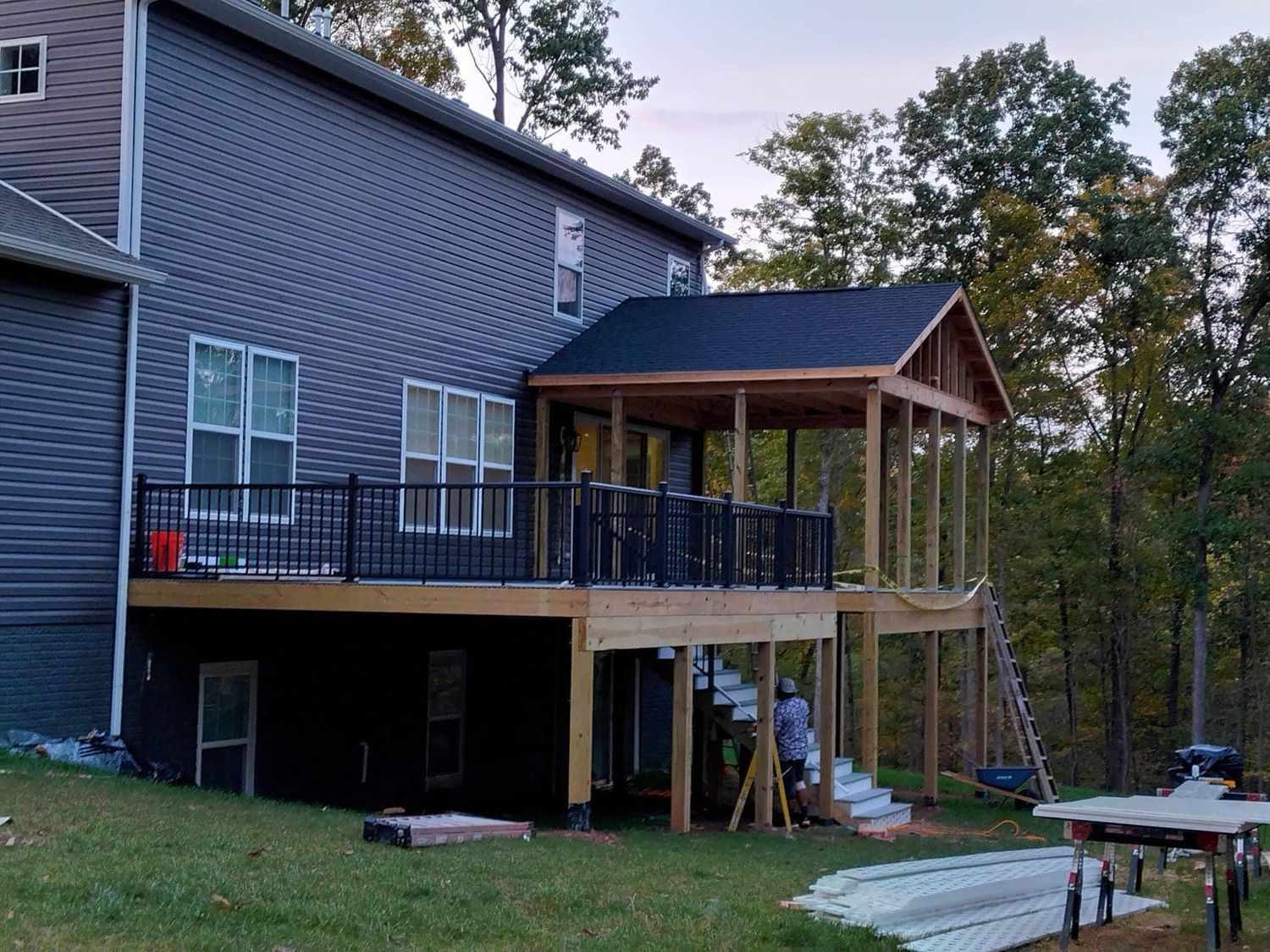 A deck and screened porch under construction on a two-story house, with black railing and wooded background.