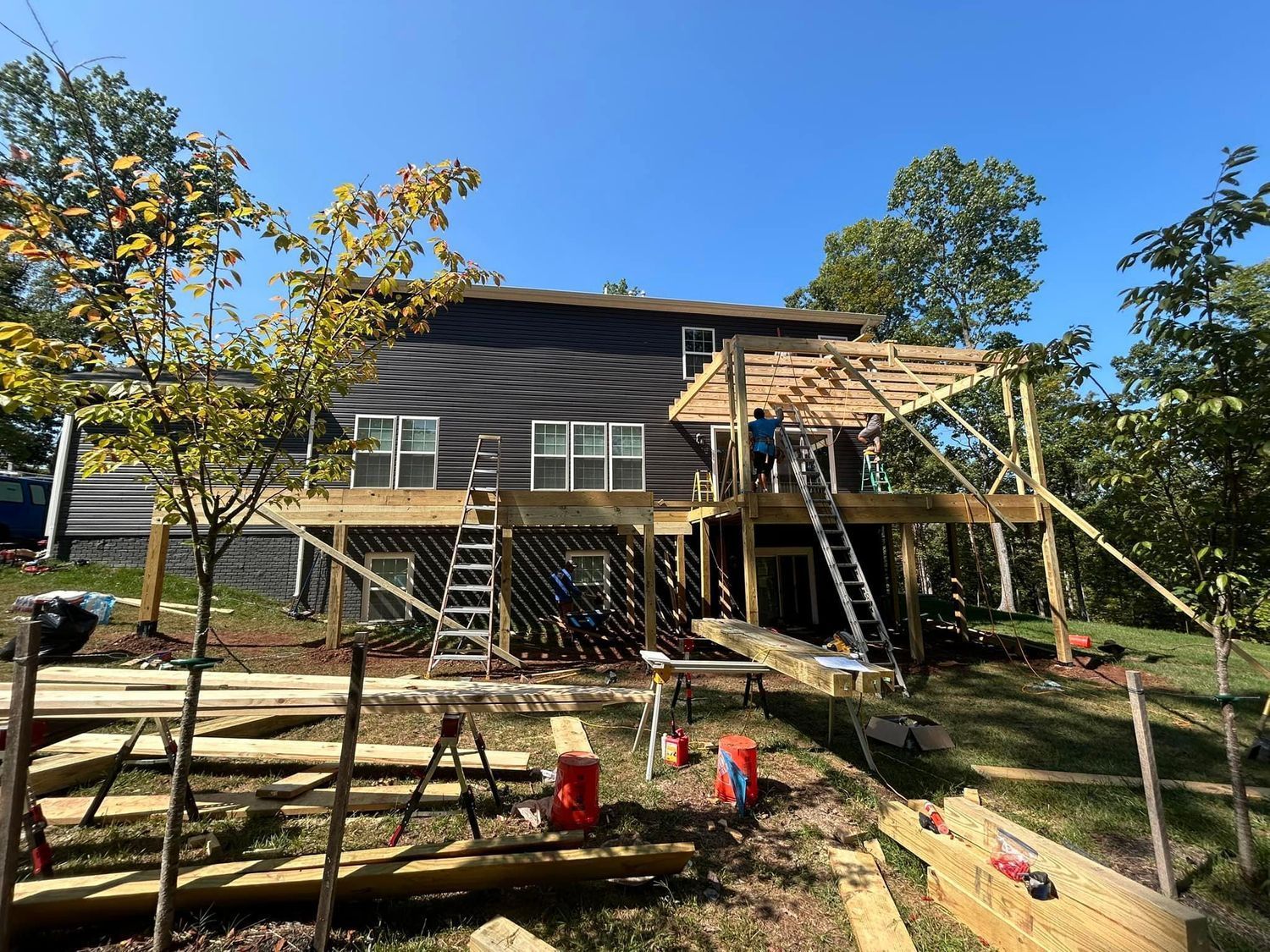 Construction of a deck addition on a dark-sided house. Workers, tools, and lumber are visible on a sunny day.
