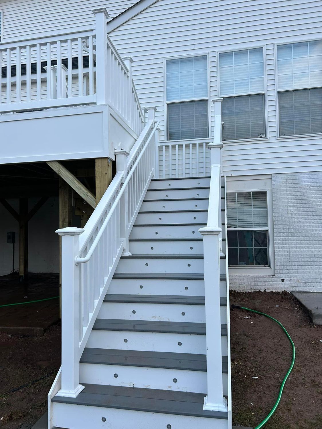 White deck stairs leading up to a house. Gray steps, white railing, green hose.