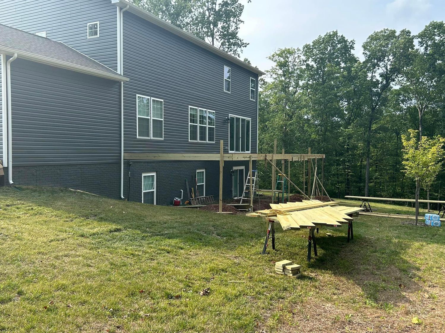 Back of a two-story gray house with deck under construction in a grassy yard, surrounded by trees.