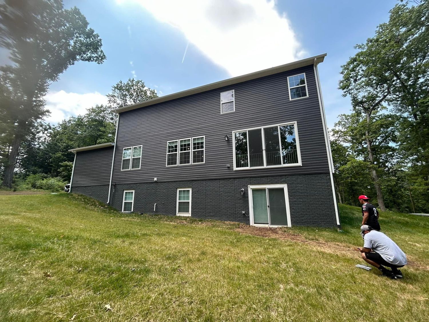 Dark gray house on a hillside with two people in front. Sunny day.
