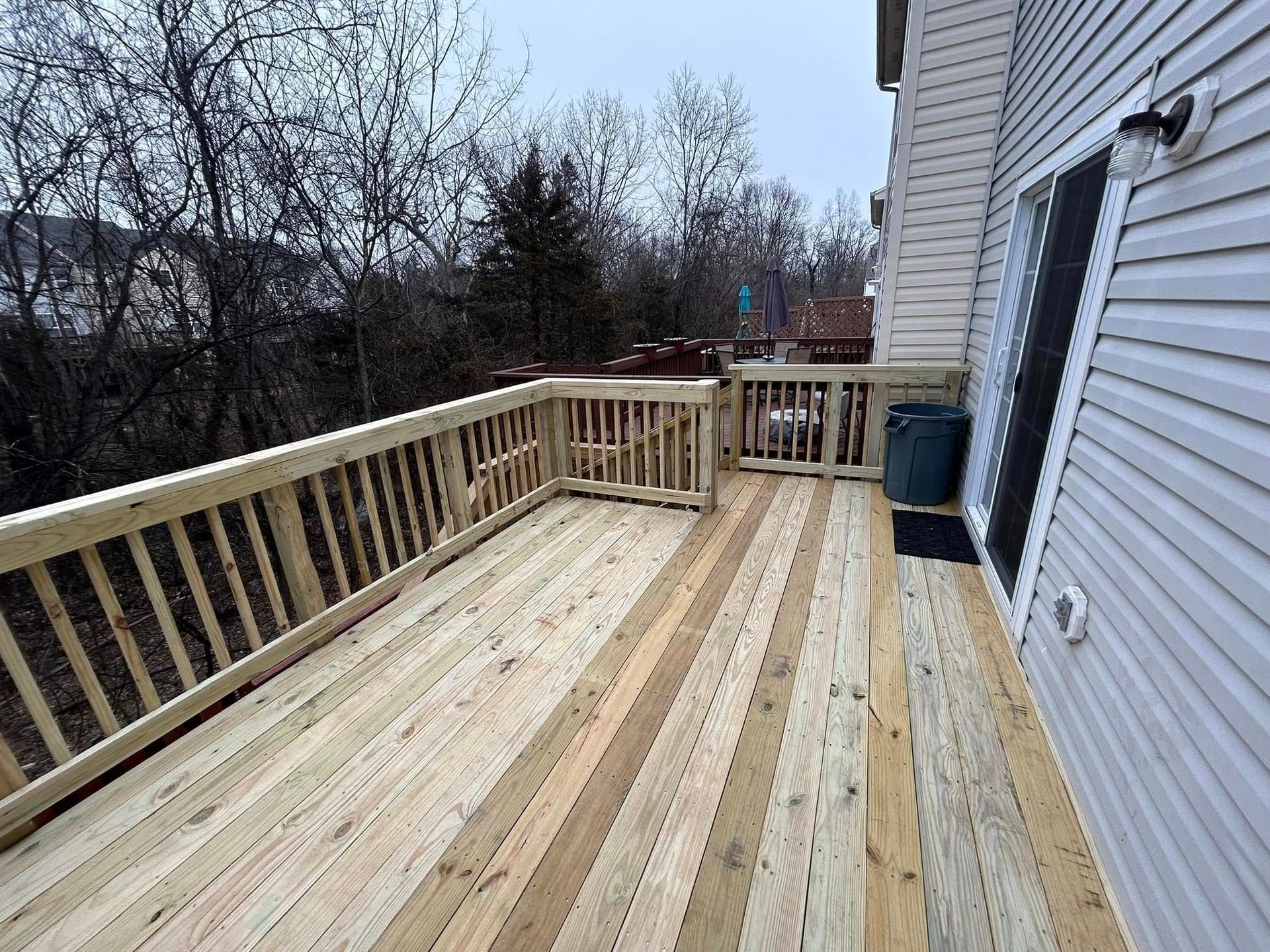 Wooden deck with railing, extending from a white-sided house. Trees in the background.