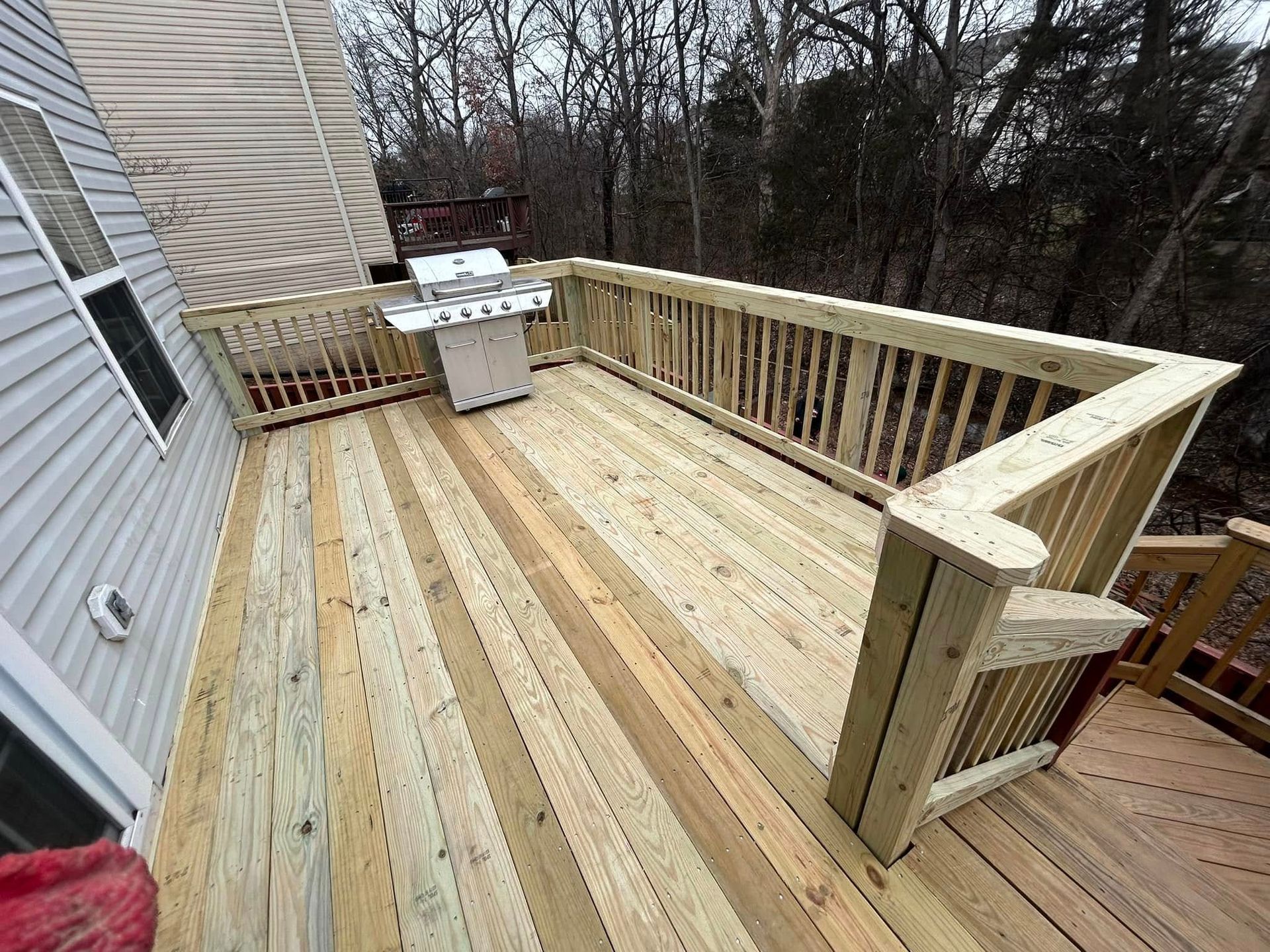 Wooden deck with grill, surrounded by railings, next to a house and trees.