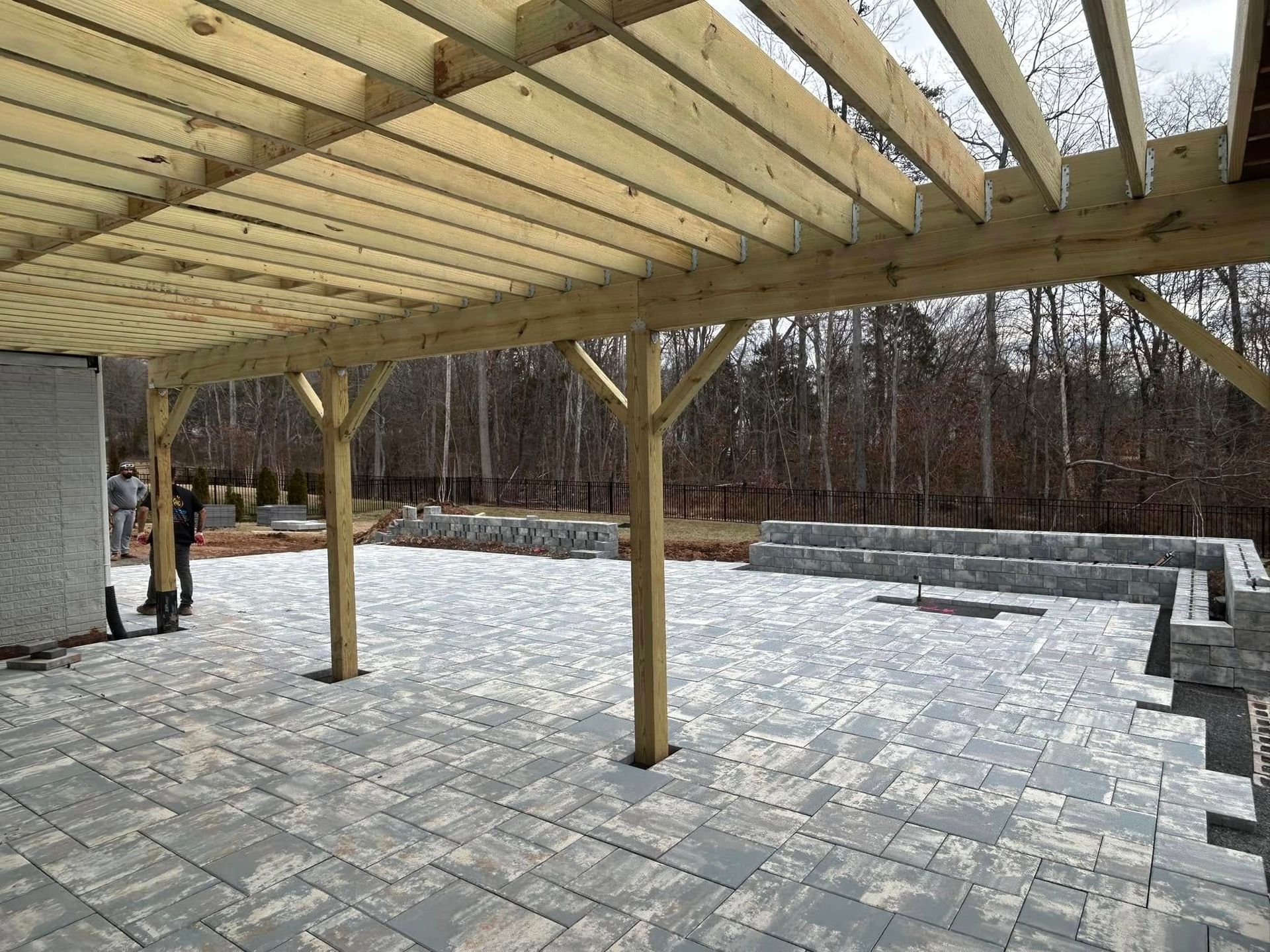 Wooden pergola over a gray stone patio; two people stand nearby; trees in the background.