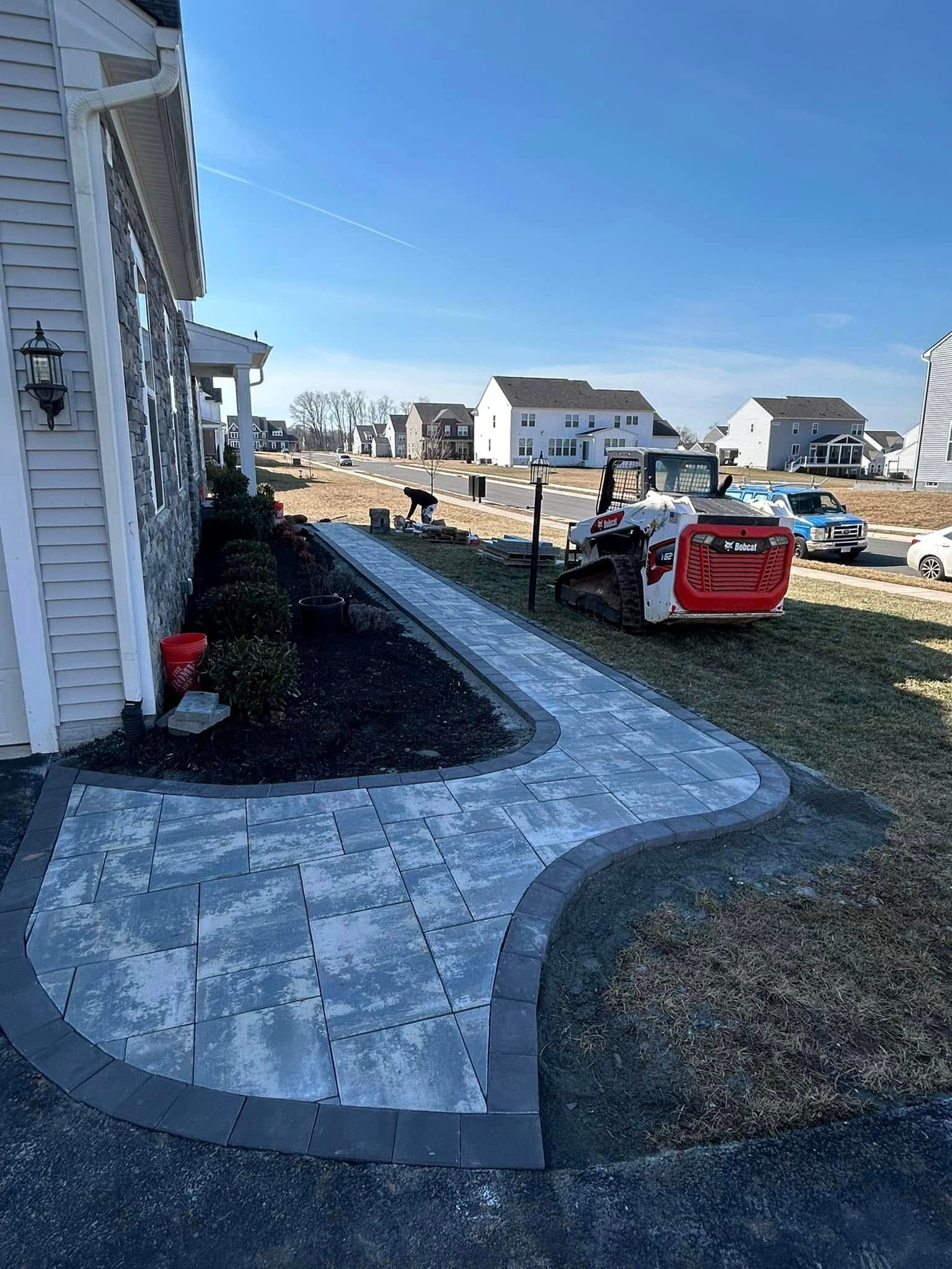 A brick pathway winds between a building and a lawn, with construction equipment nearby.