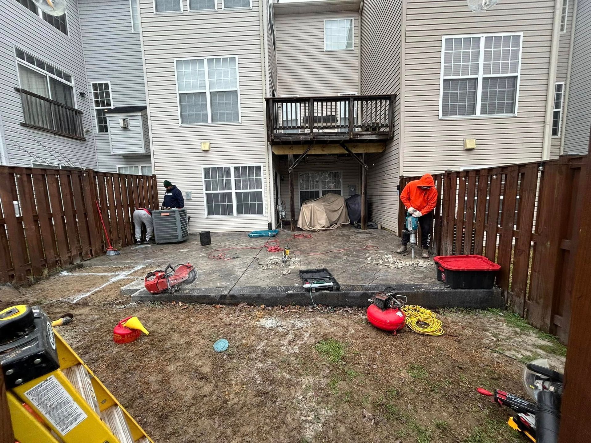 Workers in a fenced backyard remove a concrete patio. One man in an orange hoodie uses a jackhammer.