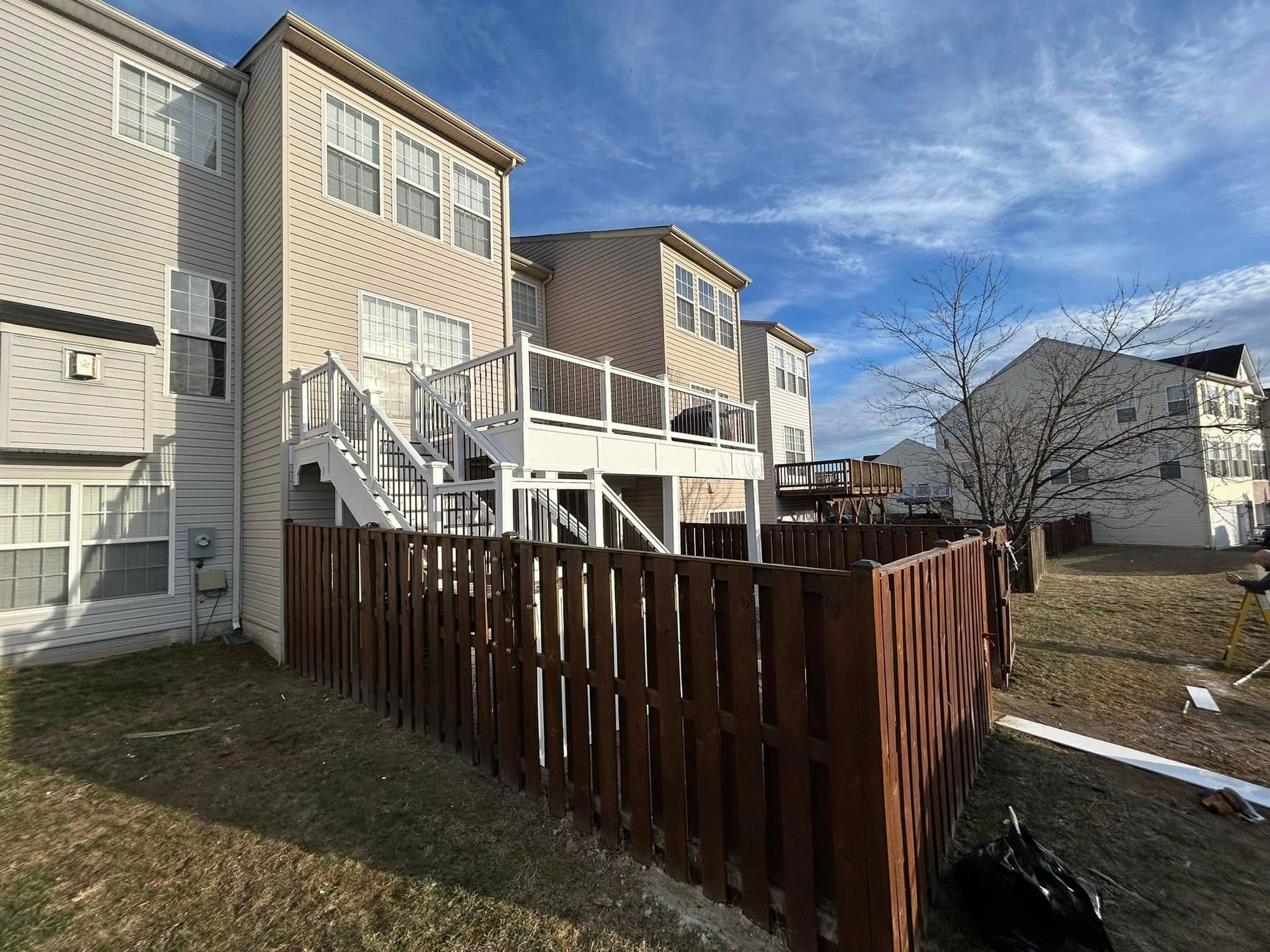Backyard with a brown wooden fence, deck, and multi-story townhouses under a blue sky.