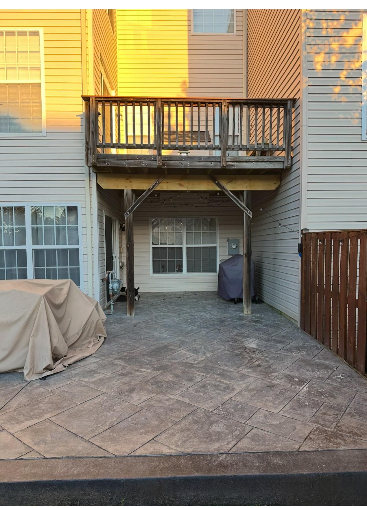 Backyard patio under a wooden deck. Grey textured concrete, beige siding, brown wooden fence.