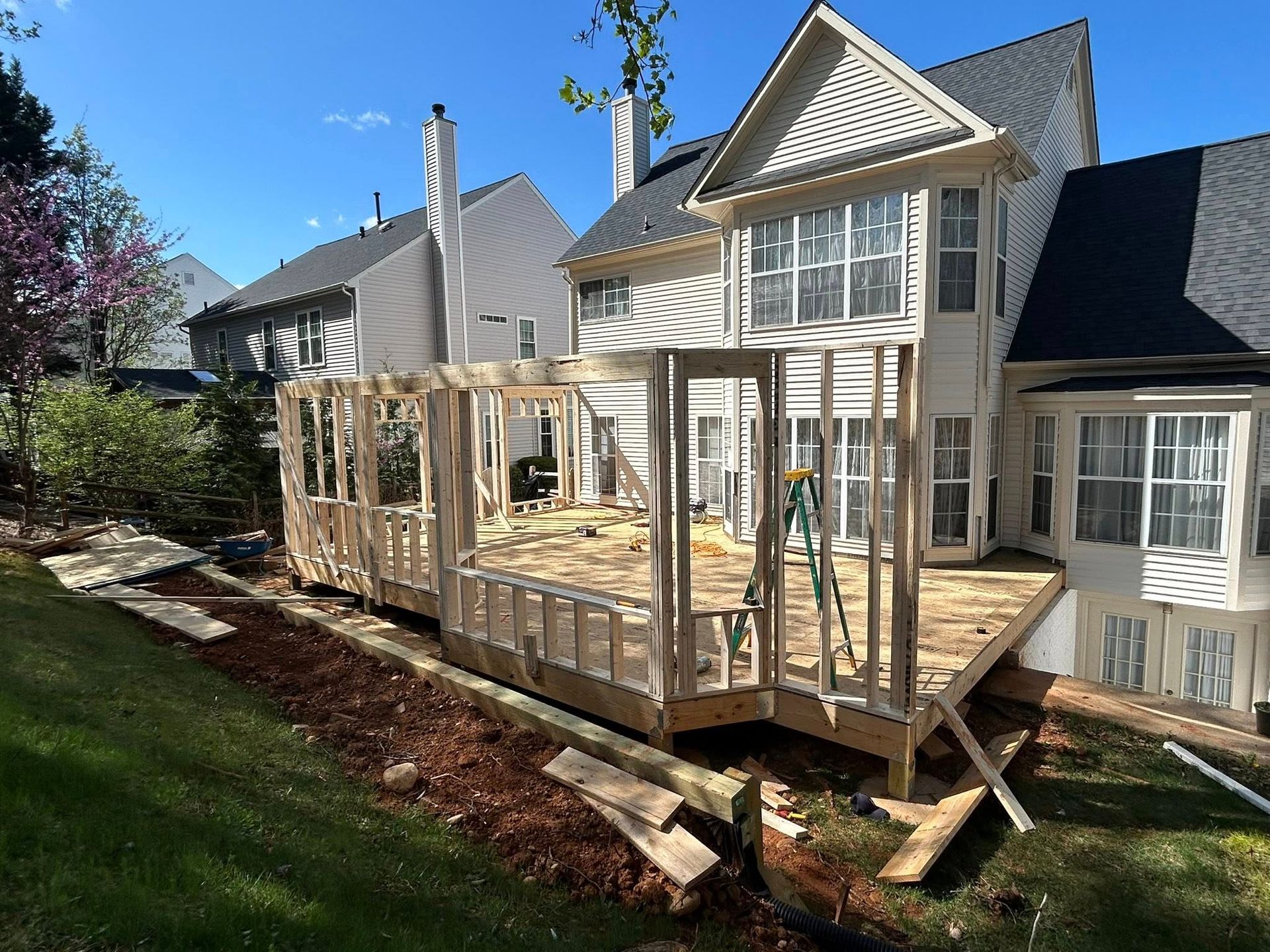 Deck under construction attached to a two-story house on a grassy hill; blue sky.