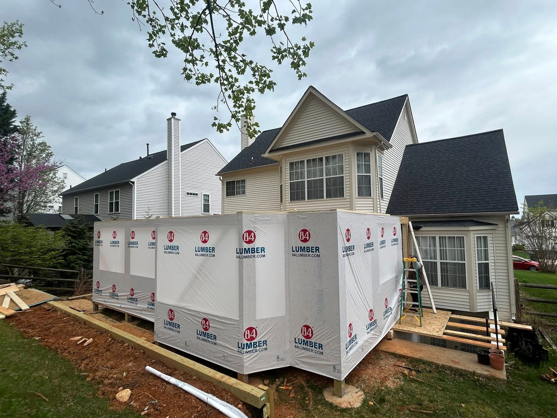 Deck construction in progress behind a two-story house; wrapped in building paper on a cloudy day.