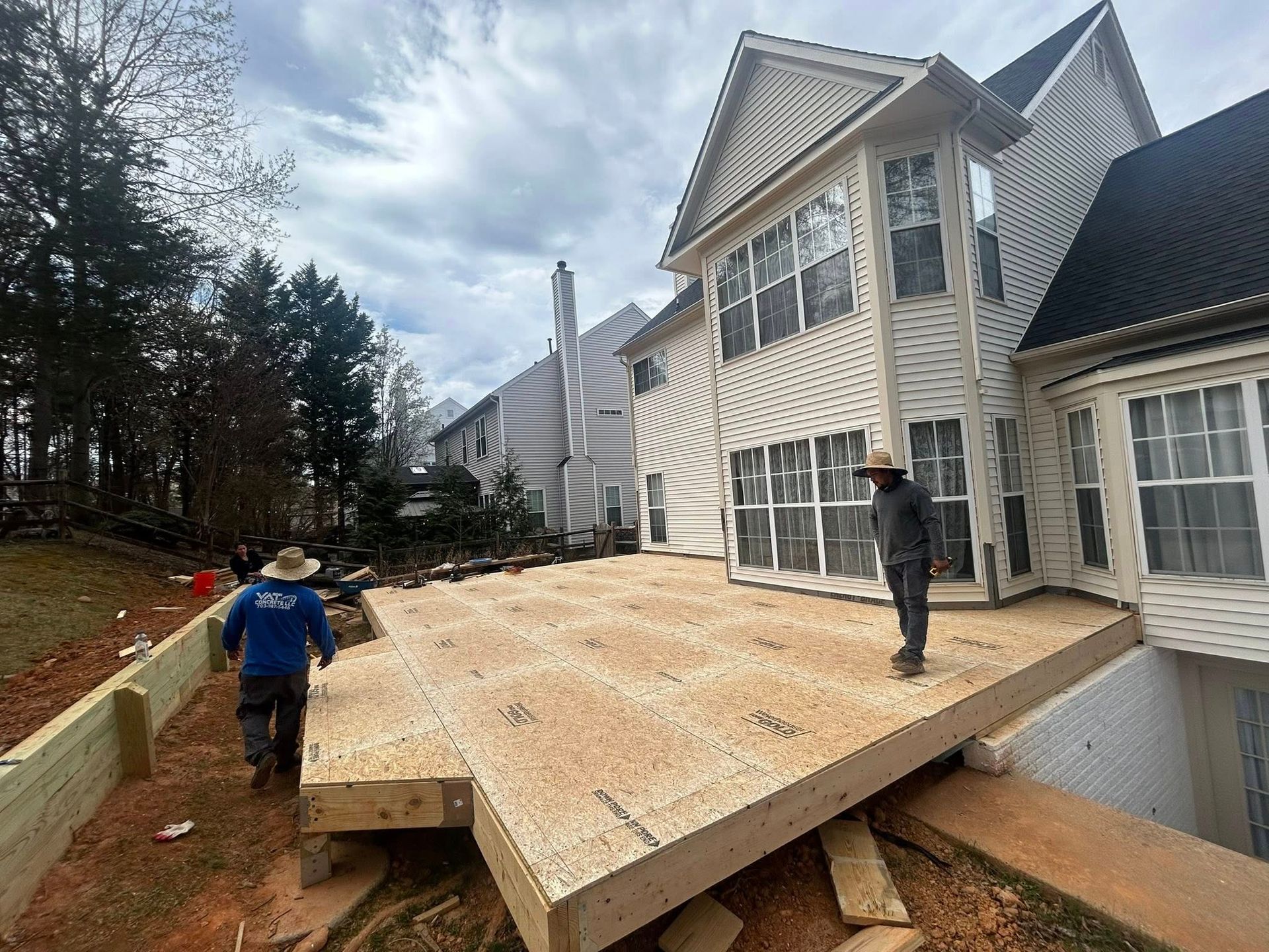 Construction workers building a wooden deck adjacent to a two-story beige house on a cloudy day.