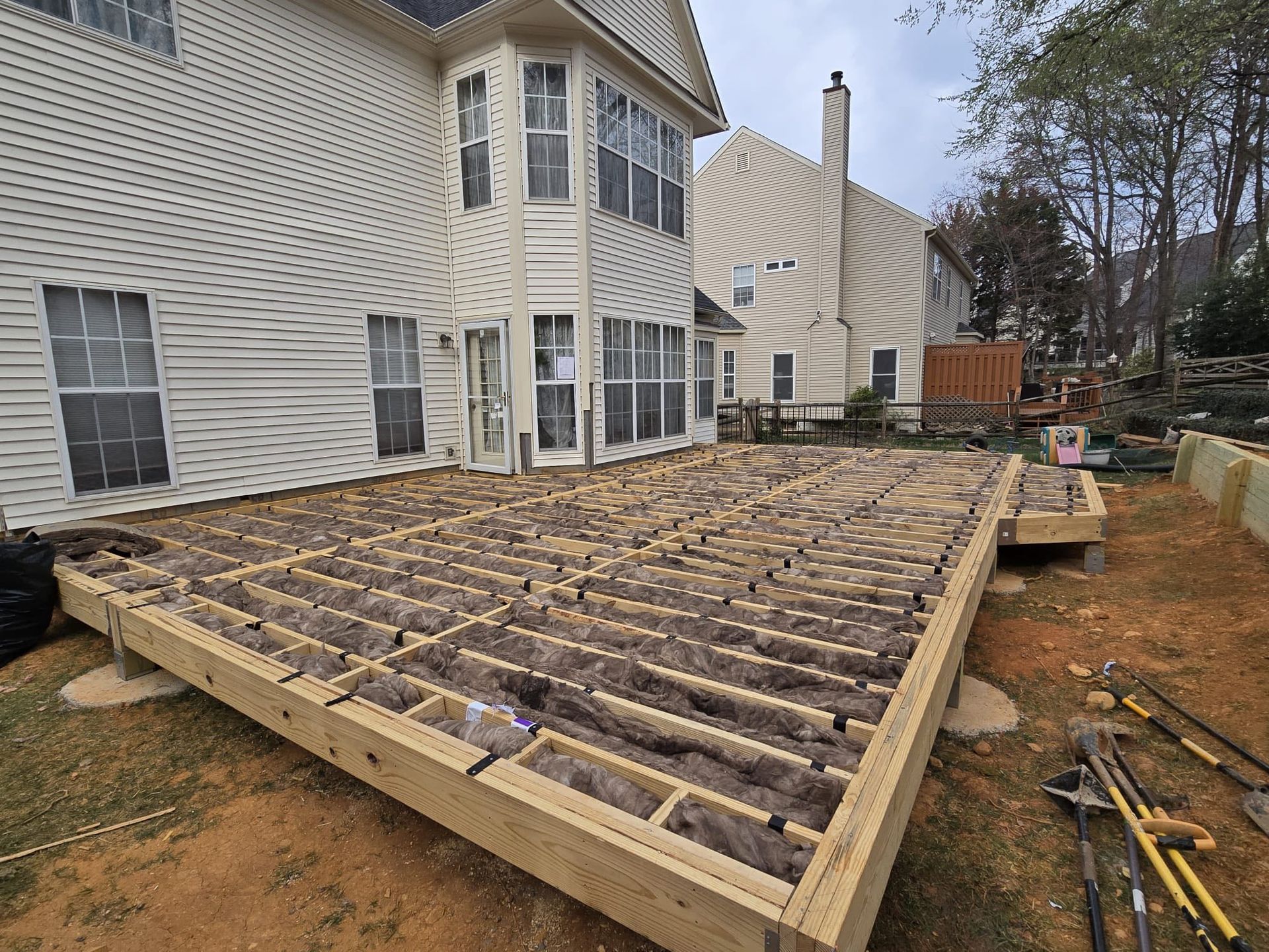Wooden deck frame under construction outside a house. Insulation visible between joists on the ground.