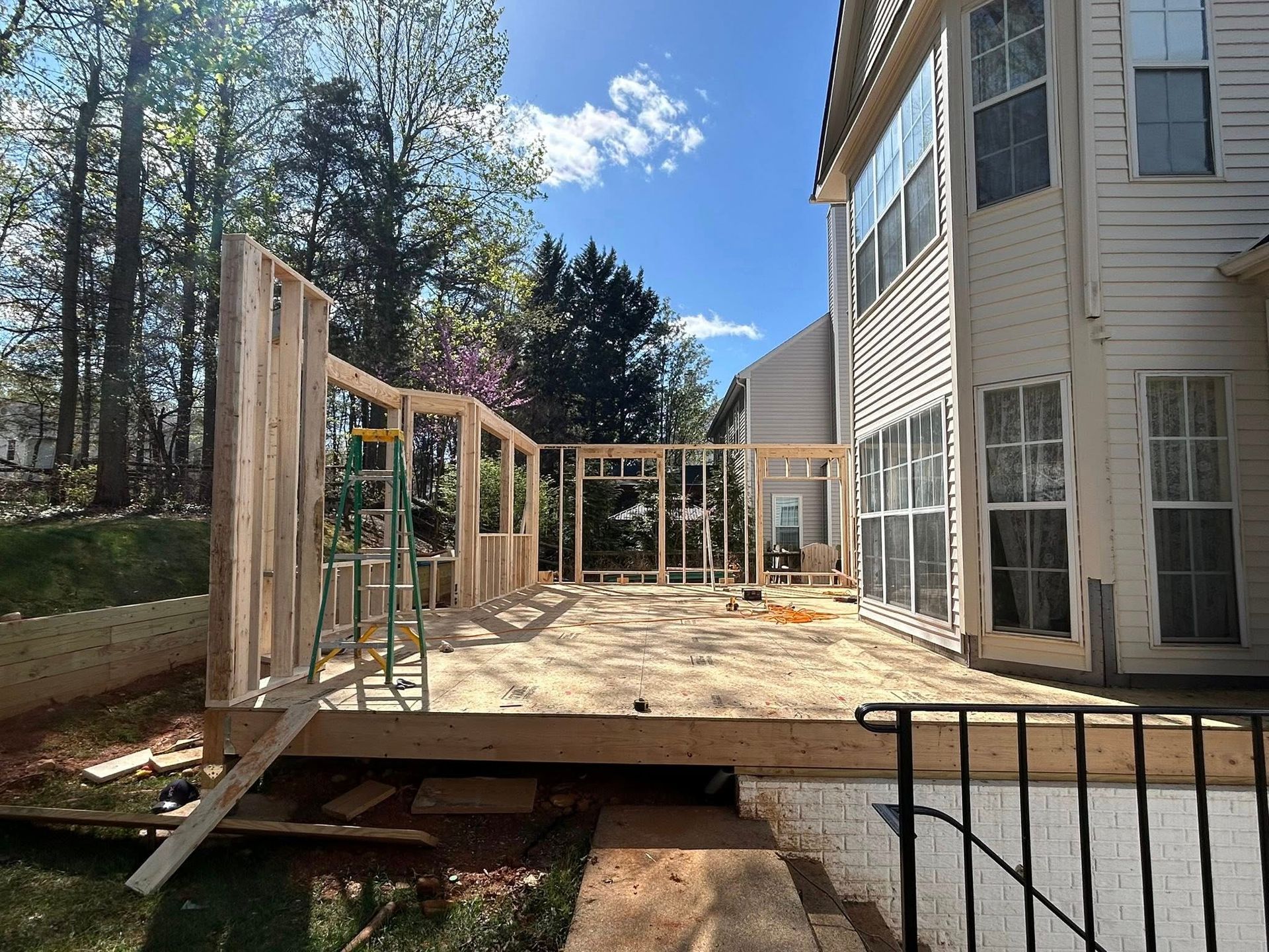 Wood-framed walls under construction on a deck next to a two-story house; sunny day.