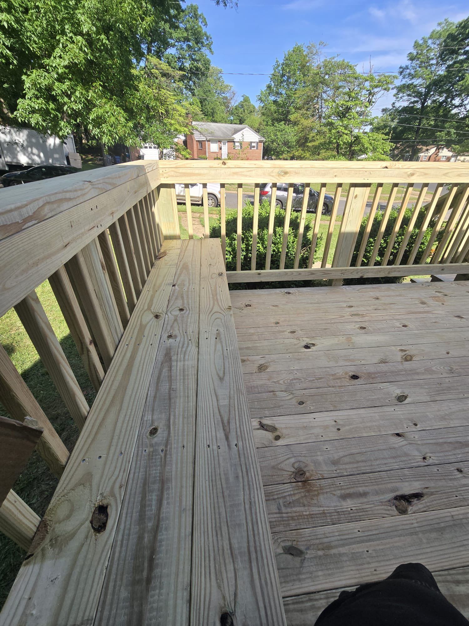 Wooden deck with railing and built-in bench, overlooking green foliage and a distant house.