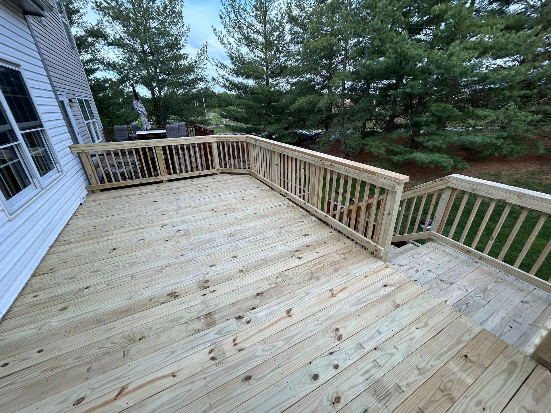 Wooden deck with railing attached to a white house, with a grassy area and trees visible.