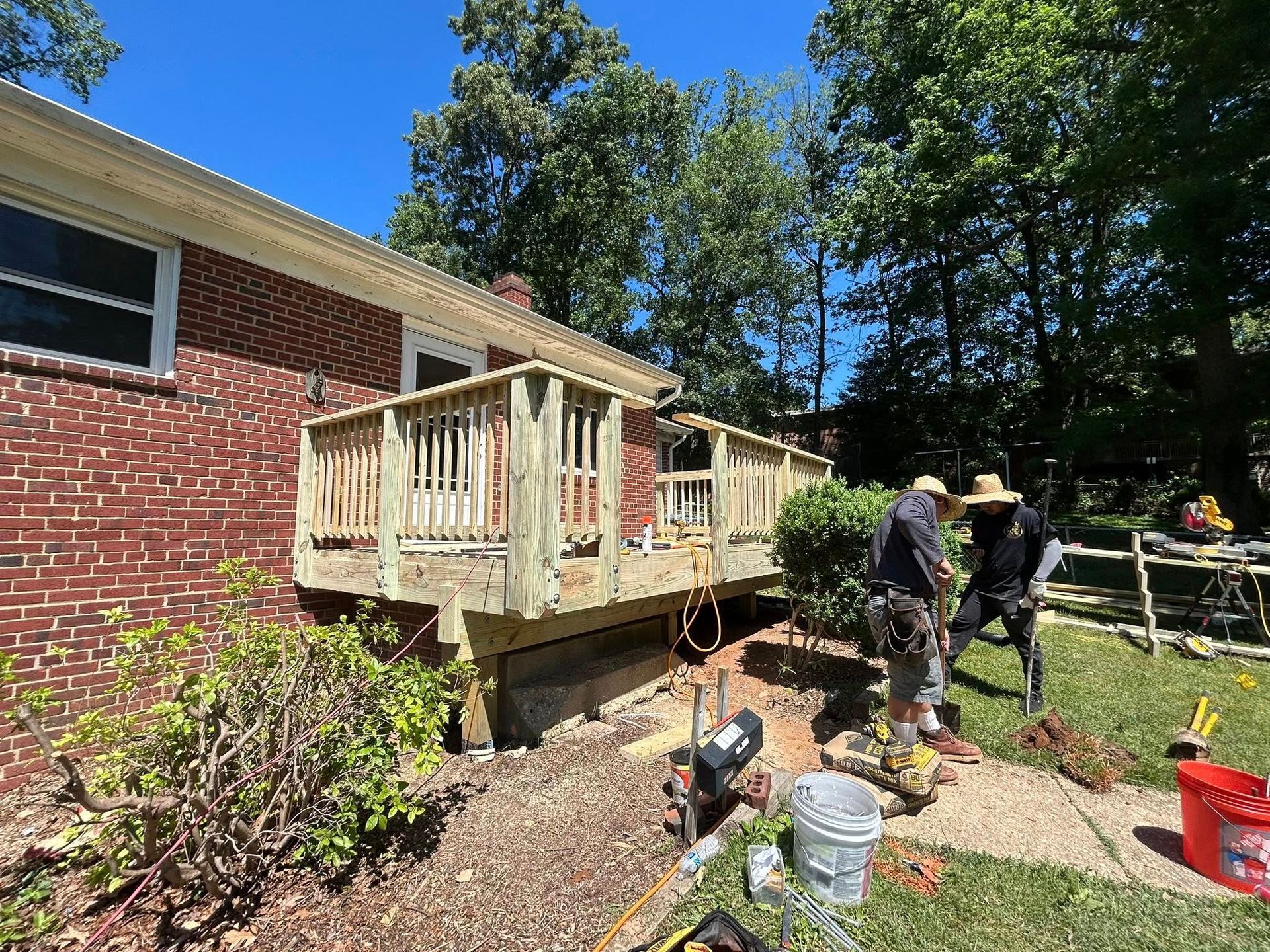 Two workers rebuild a wooden deck attached to a brick house on a sunny day.