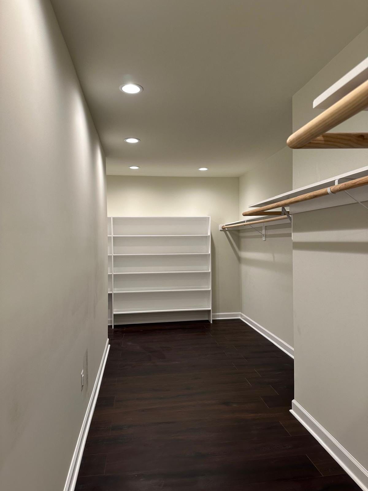 Narrow walk-in closet with dark wood floor, white shelves, and hanging rods; neutral walls and ceiling.