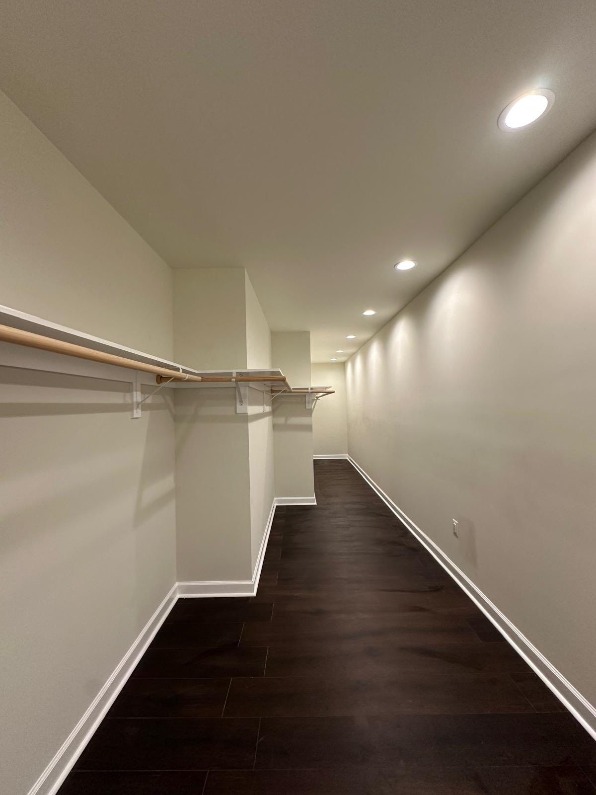 Narrow walk-in closet with dark wood floor, white walls, a clothes rod, and recessed lighting.