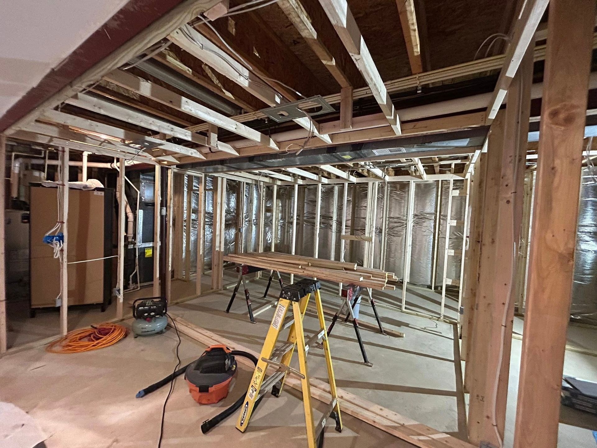 Construction site, interior of a room. Wooden framing and insulation visible. Tools and debris on the floor.