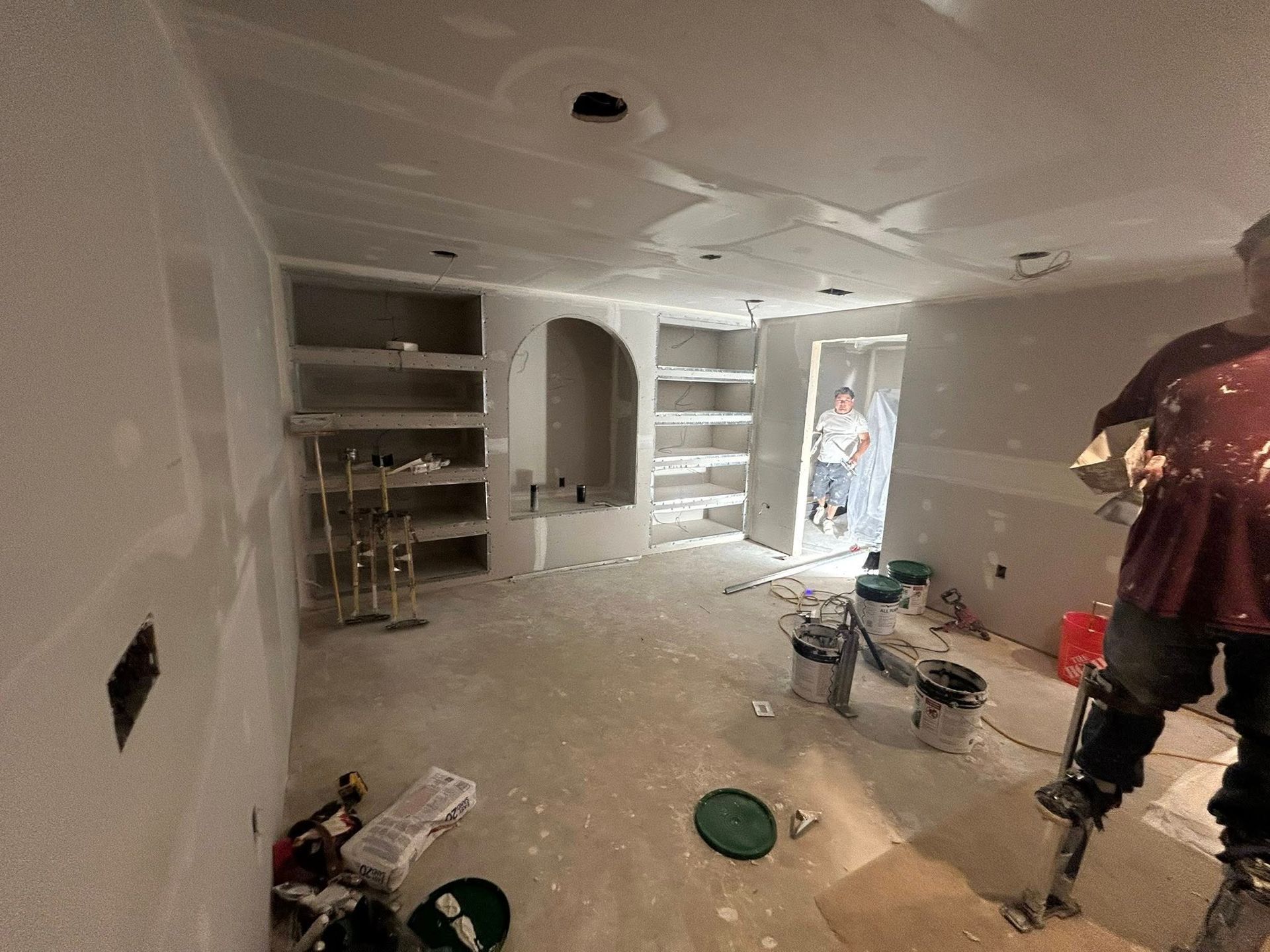 A person on stilts finishing drywall in a room with built-in shelves, another person in a doorway.