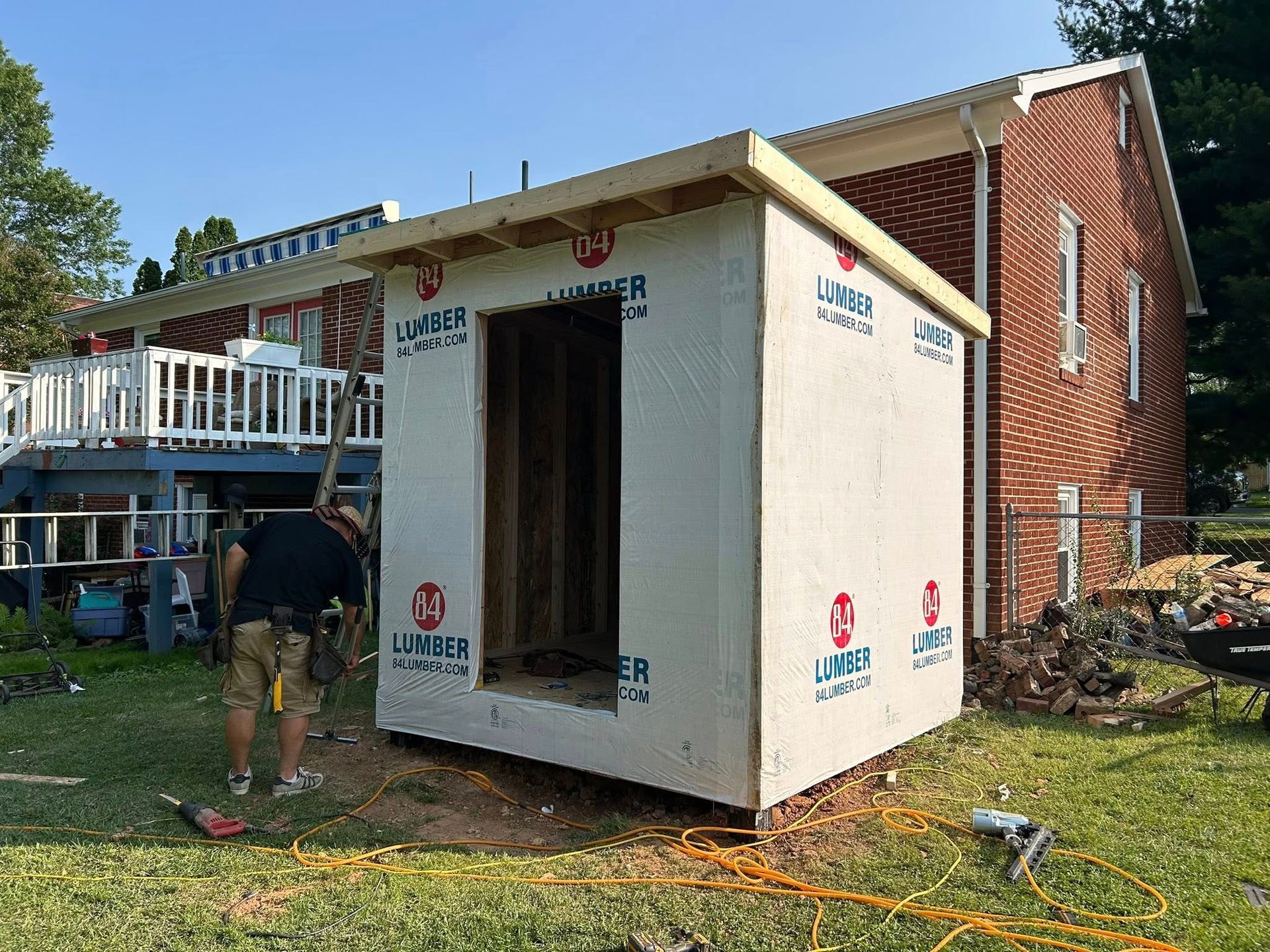 A man builds a small shed next to a brick house, applying lumber; sunny outdoors.