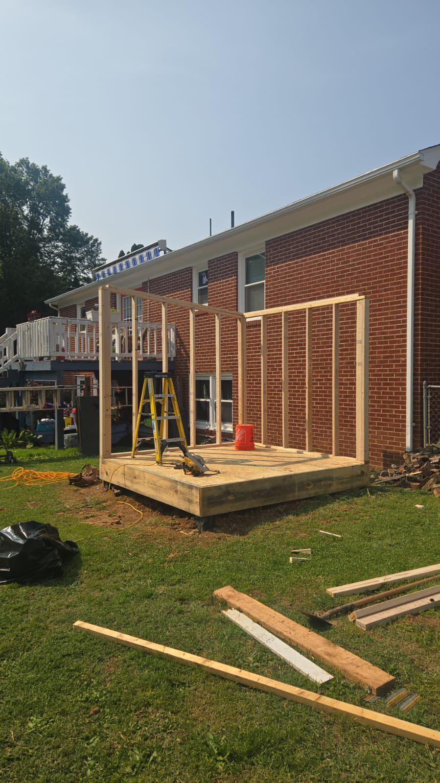 Wooden structure under construction against a brick house, with lumber and tools on the ground.