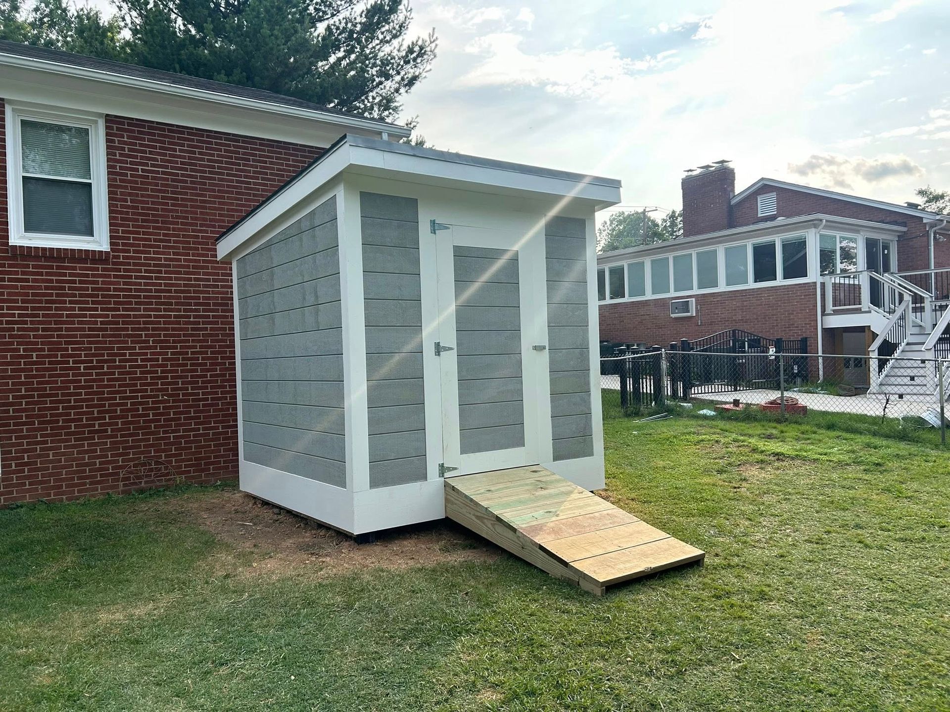 A small, white and gray shed with a wooden ramp sits in a grassy yard, next to a brick building.