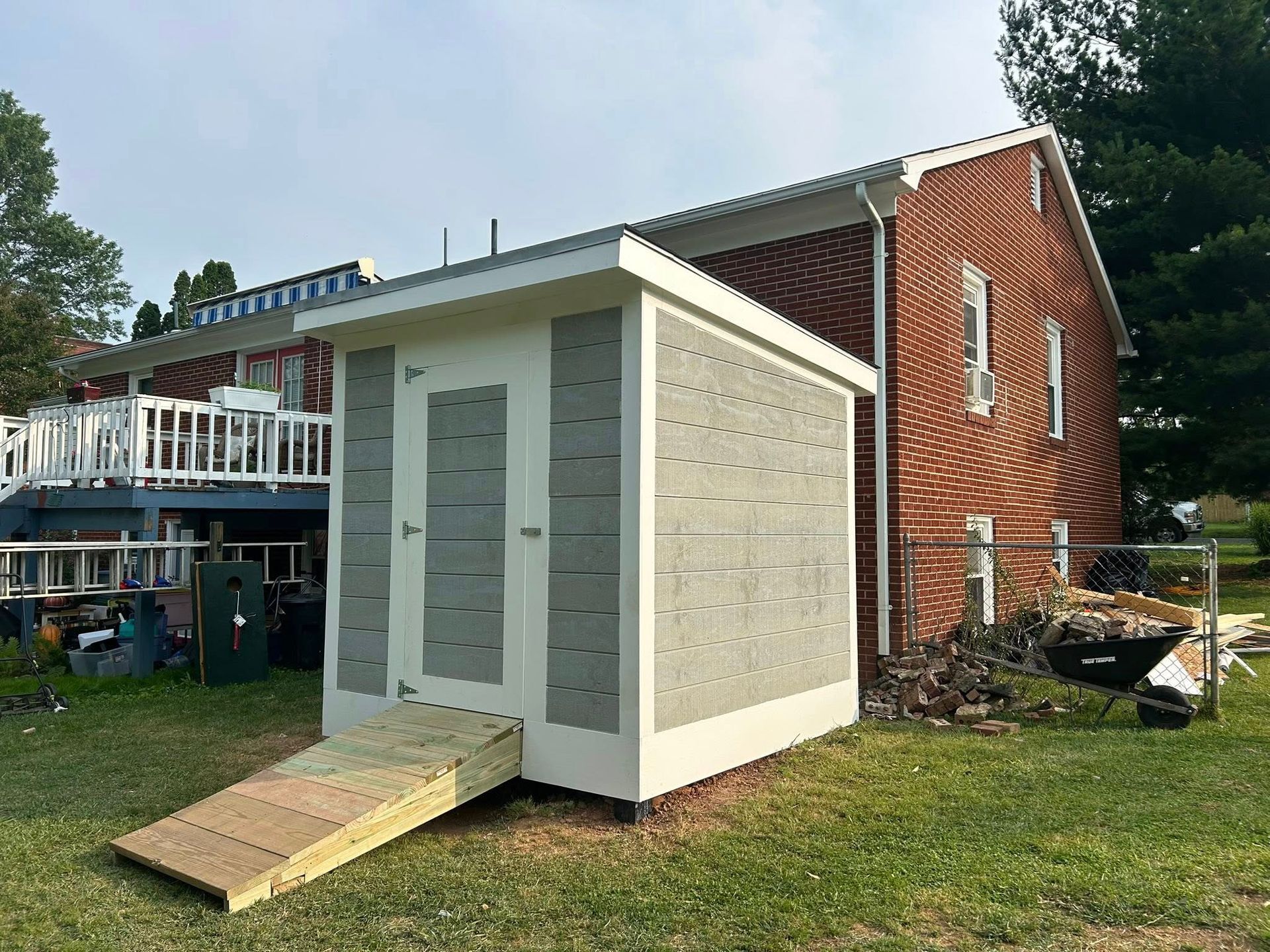 A small white and gray shed with a wooden ramp next to a brick house.
