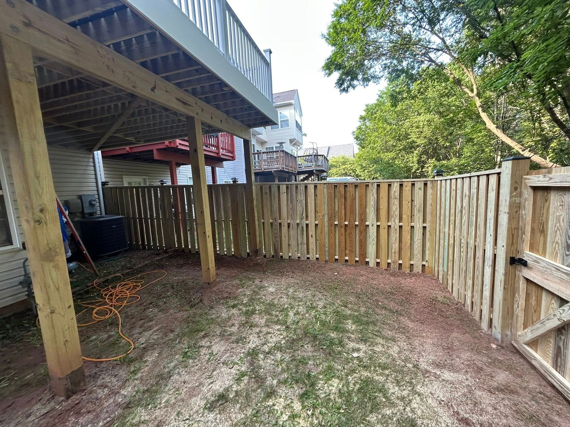 Wooden fence encloses a backyard beneath a deck. Dirt and grass ground.
