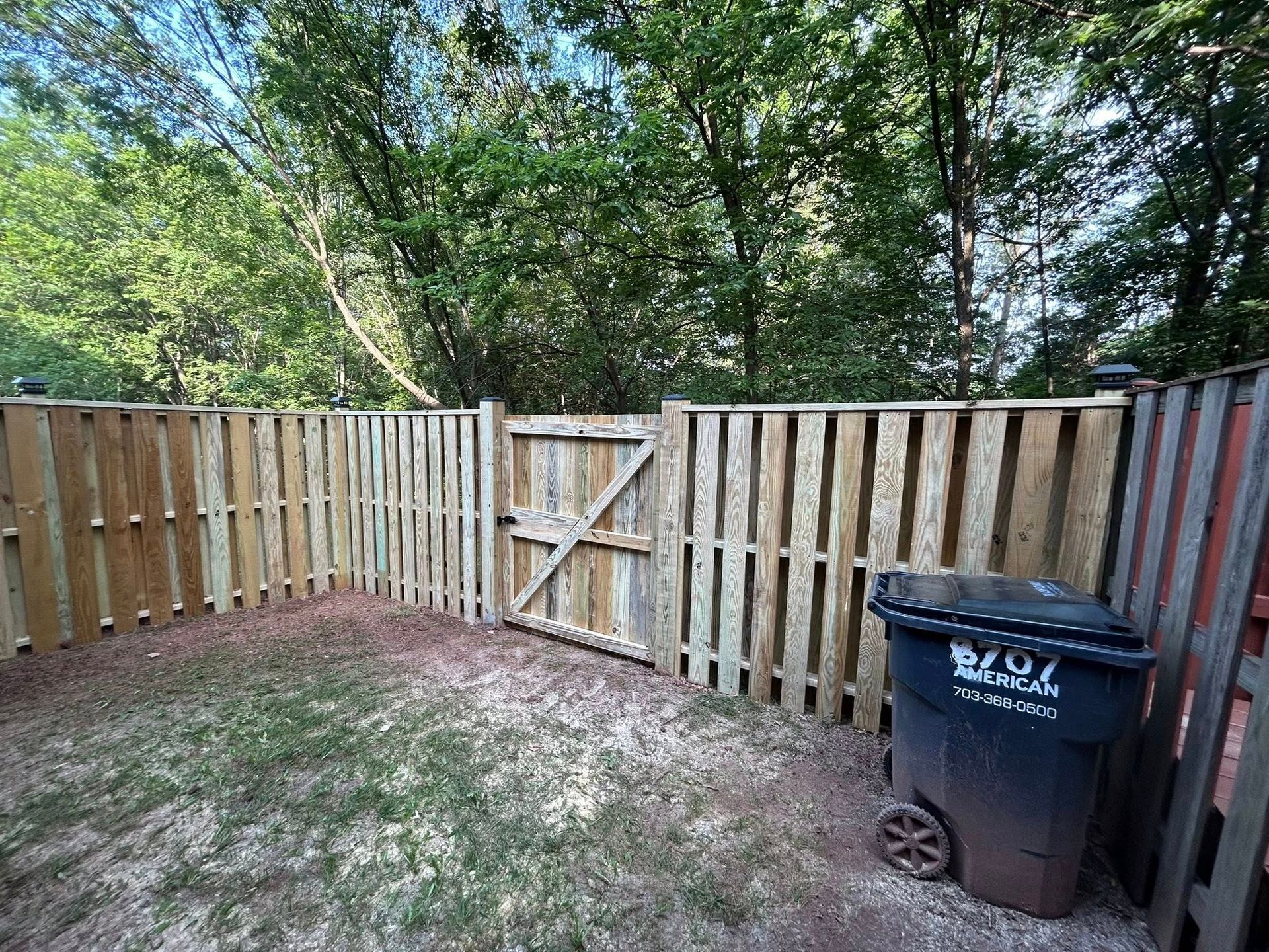 Wooden fence with gate, enclosing a small yard, next to a trash can and greenery.