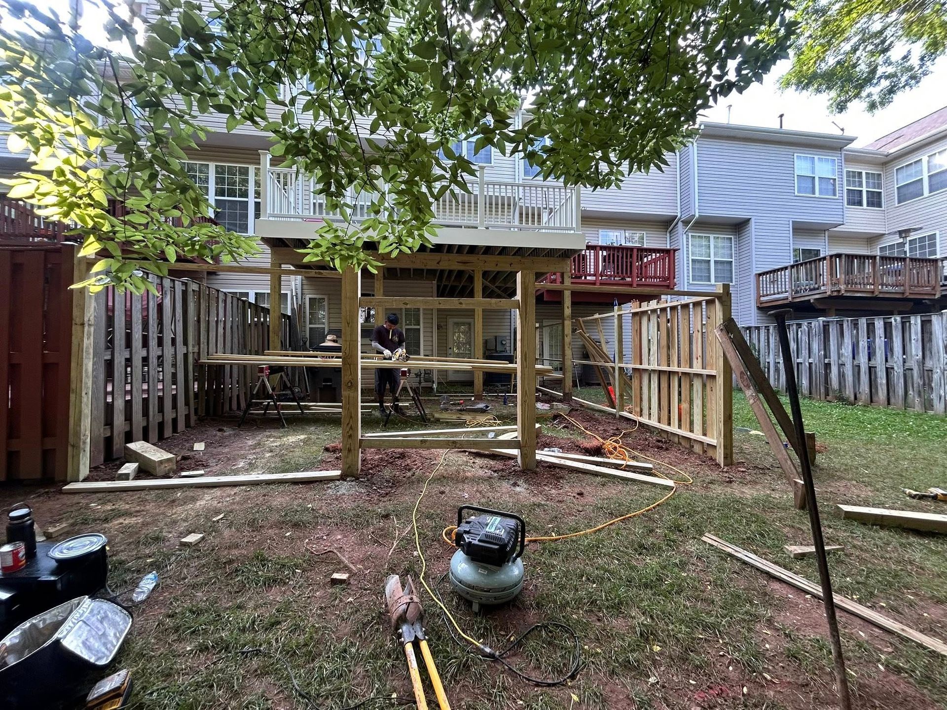 Backyard construction: worker building a wooden structure under a deck, surrounded by tools and fencing.