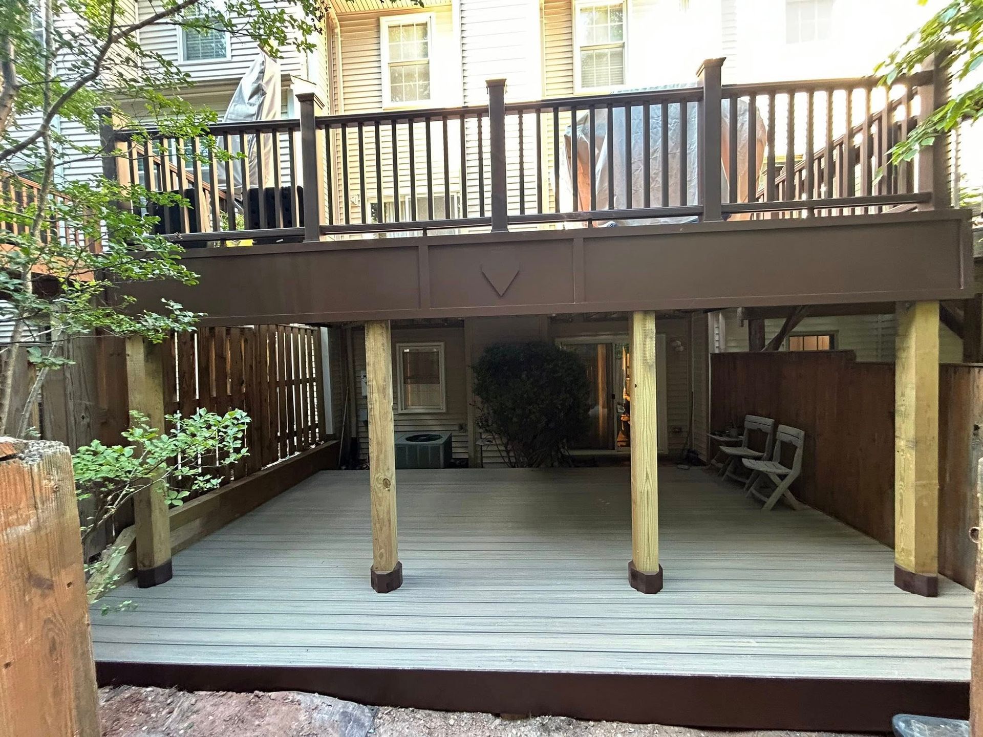 Two-level brown wooden deck in backyard. Upper deck with railing. Lower deck with pillars.