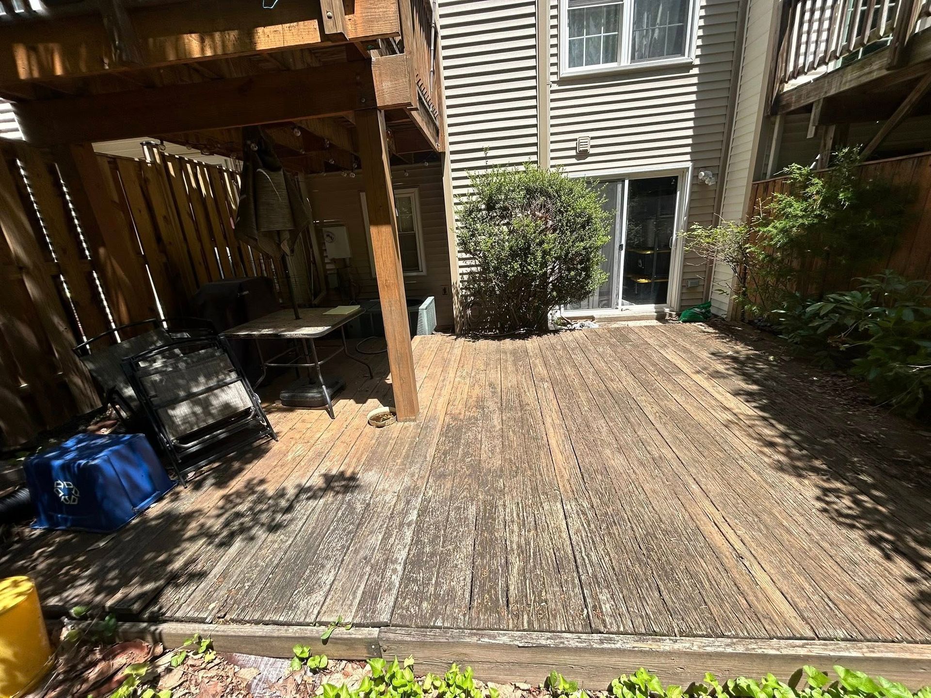 Wooden deck with weathered planks, leading to a house. Patio furniture and a bush are visible.
