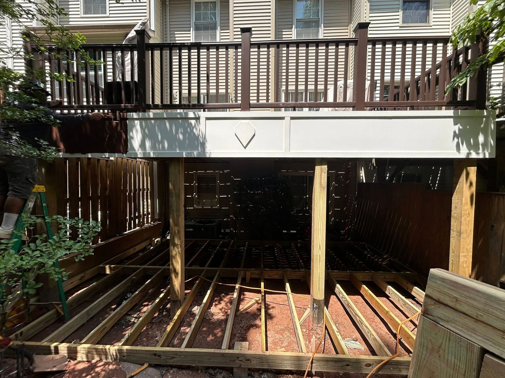 A deck under construction with exposed joists and support posts beneath an existing upper deck.