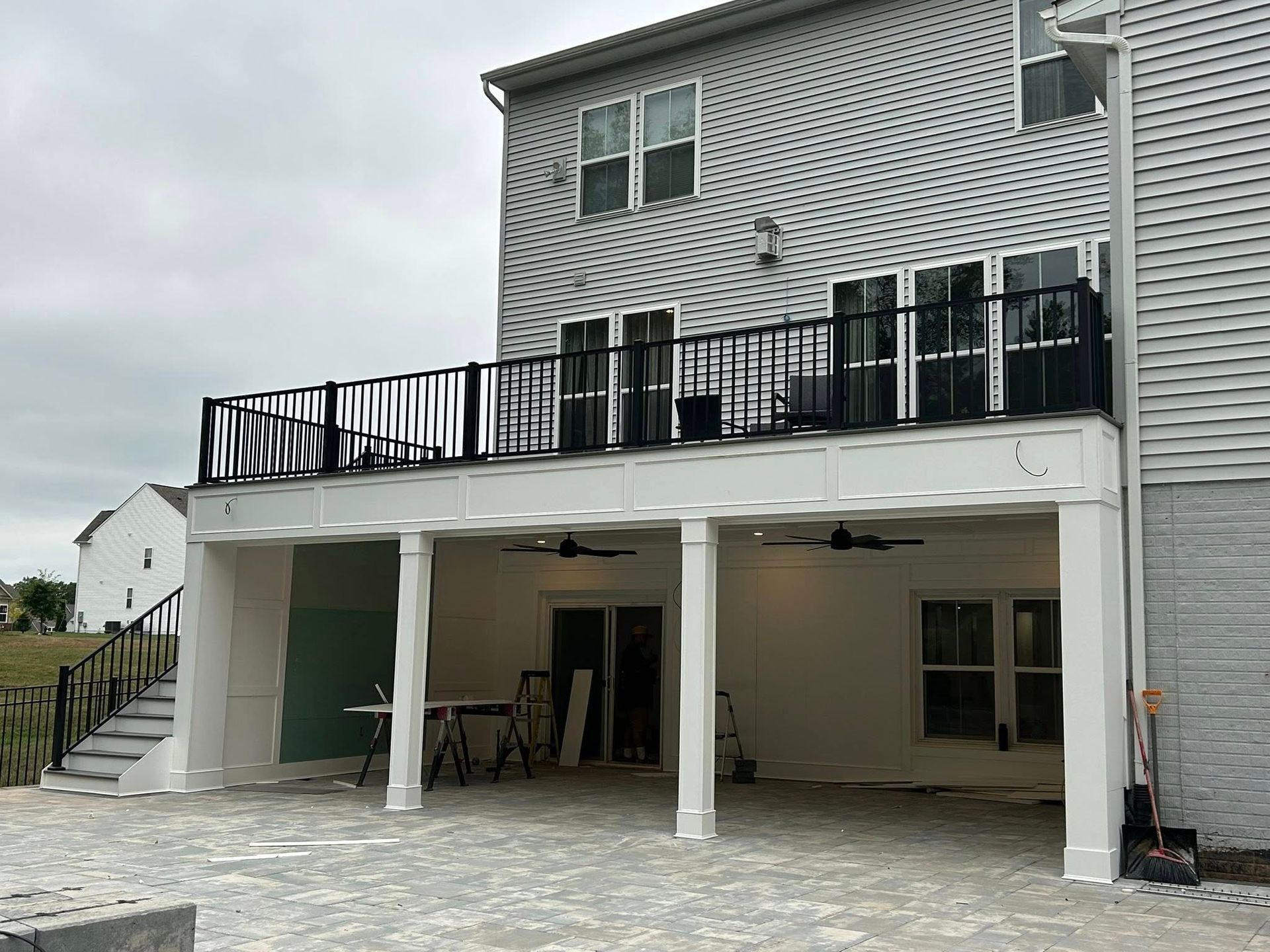 Two-story house with a deck, white pillars, and stone patio under overcast skies.
